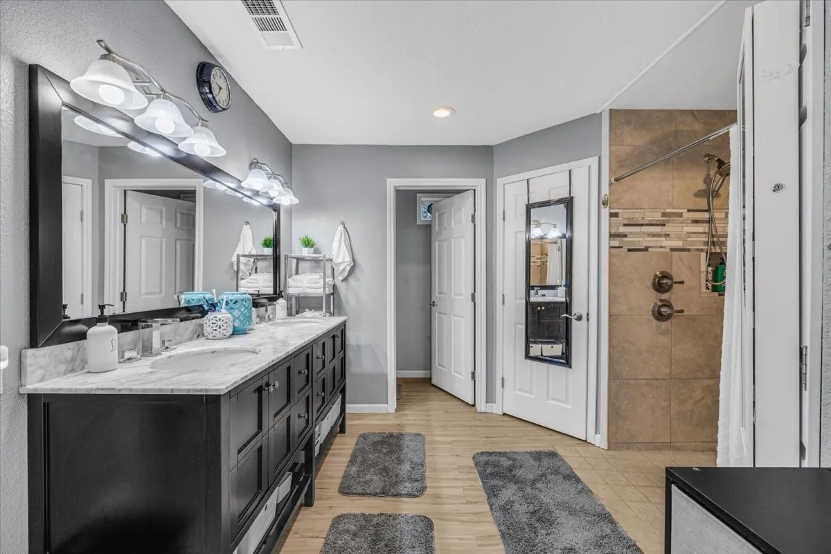Double-sink vanity featuring a stone-surface countertop, dark-finish cabinetry, a full-width mirror, integrated wall-mounted lighting, and a walk-in shower with decorative tile inlay