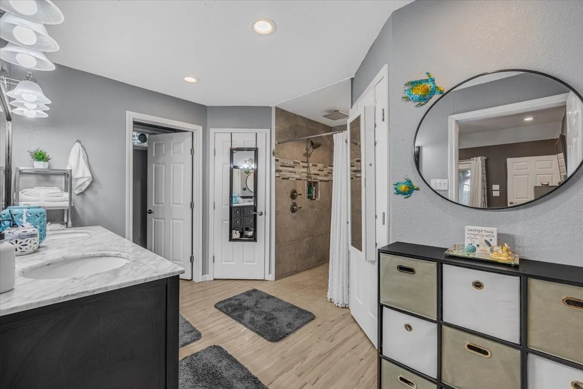 Bathroom featuring stone-finish vanity countertops with dual recessed sinks, dark cabinetry, wood-finish flooring, and a walk-in shower with tiled surround and an integrated niche