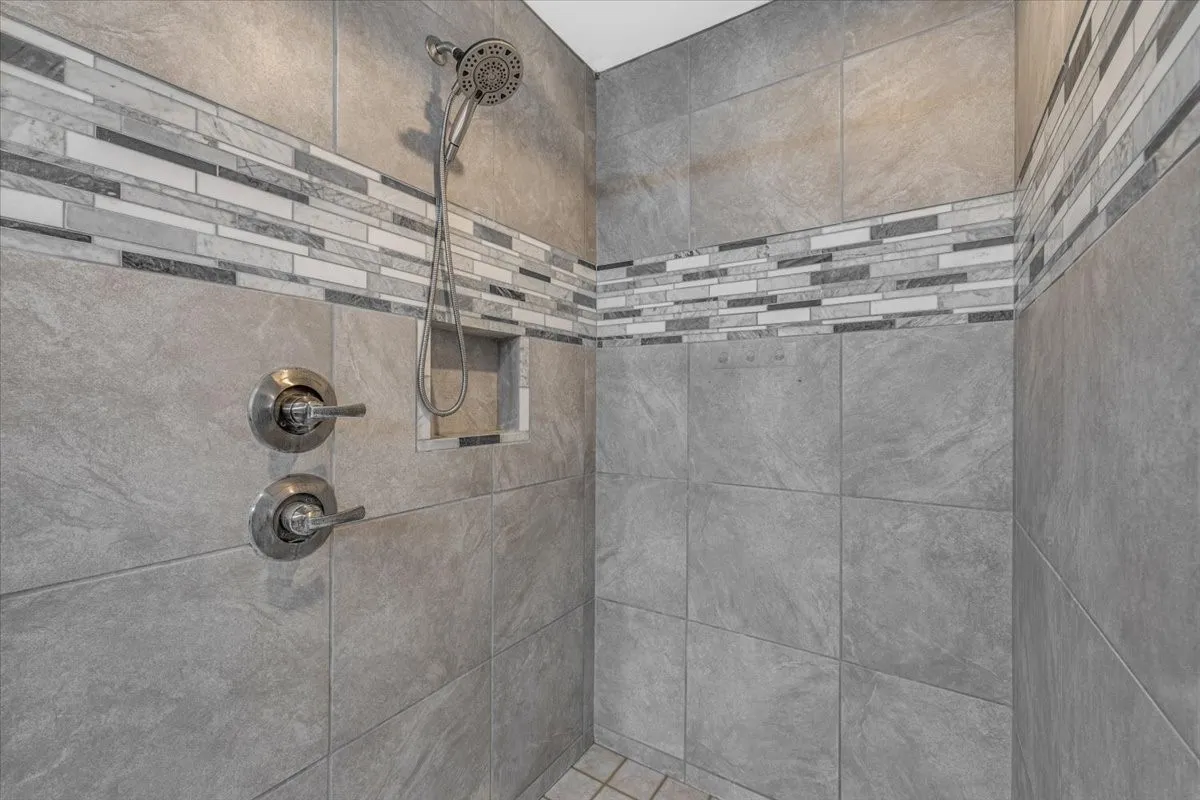 Walk-in shower featuring large-format grey tile, a decorative linear mosaic accent border, a built-in tiled niche, and chrome rainfall shower head with dual-handle controls