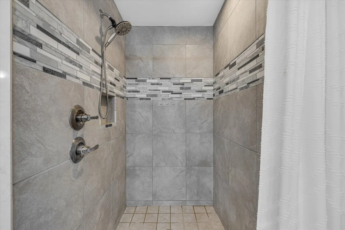 Shower stall featuring large-format grey wall tile, a horizontal mosaic tile accent border, a built-in recessed soap niche, and a wall-mounted shower head with adjustable handheld attachment