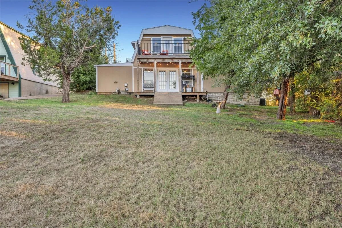 Rear exterior featuring a covered deck, second-story balcony with metal railings, French doors, and mature landscaping