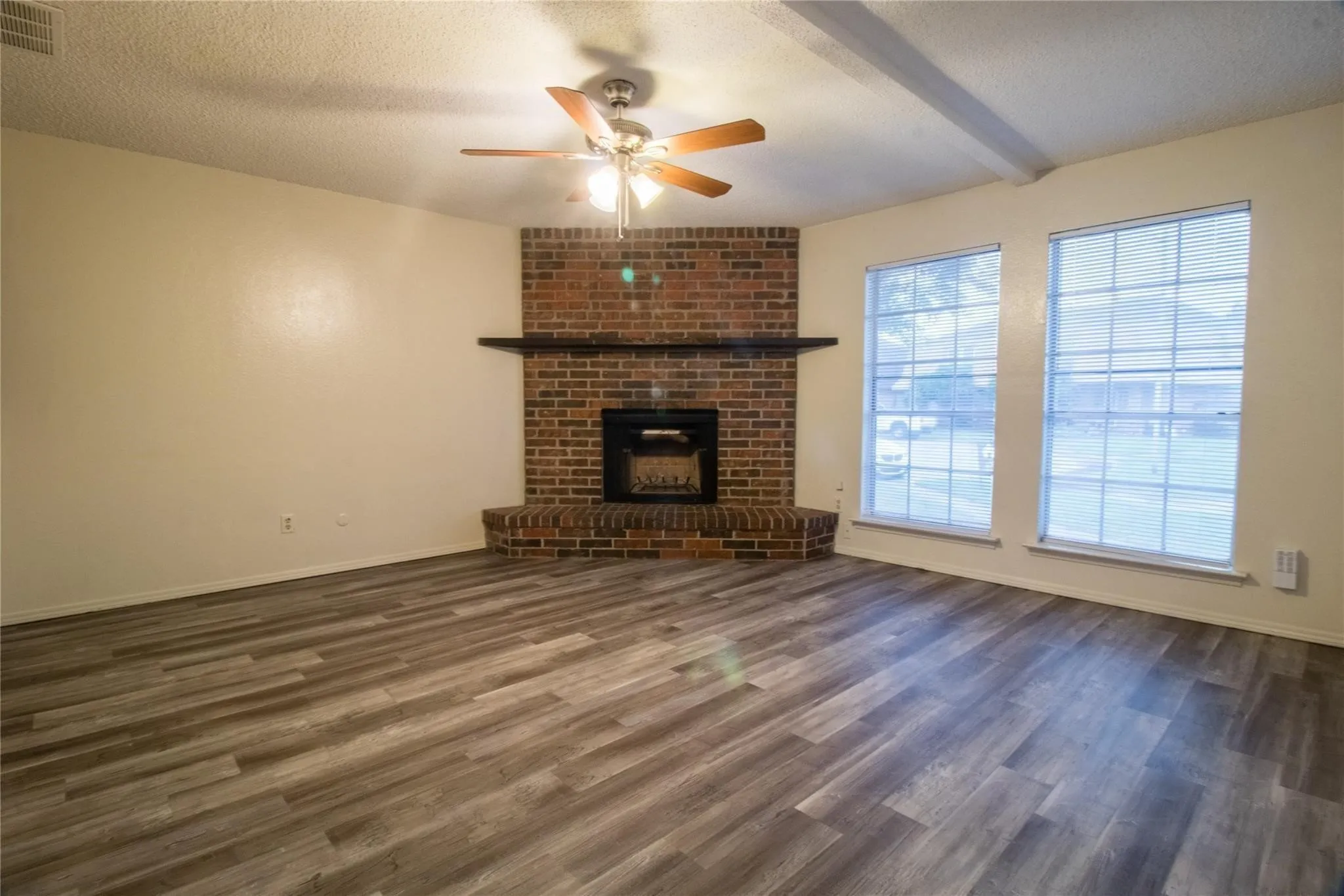 Corner brick fireplace with a wood mantel and raised hearth, wood-finish flooring, dual grid-pattern windows, and a multi-blade ceiling fan
