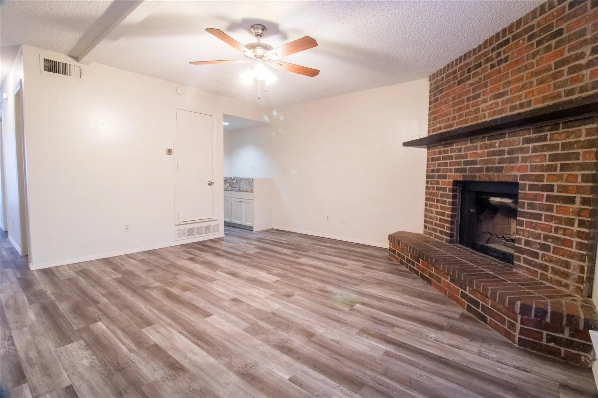 Living area featuring wood-finish flooring, floor-to-ceiling brick fireplace with a raised hearth, wood mantel, and ceiling fan with integrated lighting