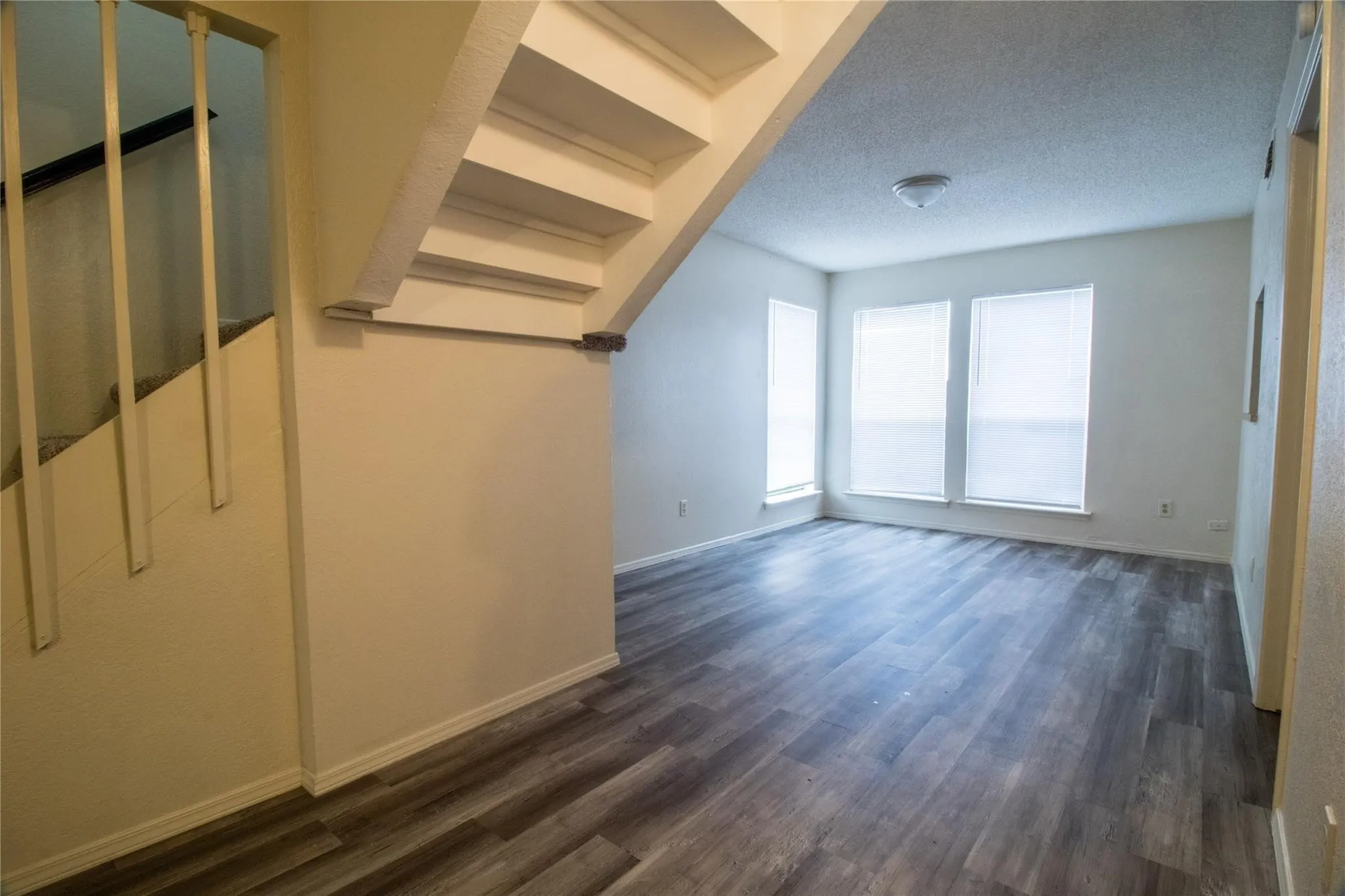 Open floor plan featuring wood-finish flooring, staircase with vertical balusters, dual windows with integrated blinds, and flush-mount ceiling lighting