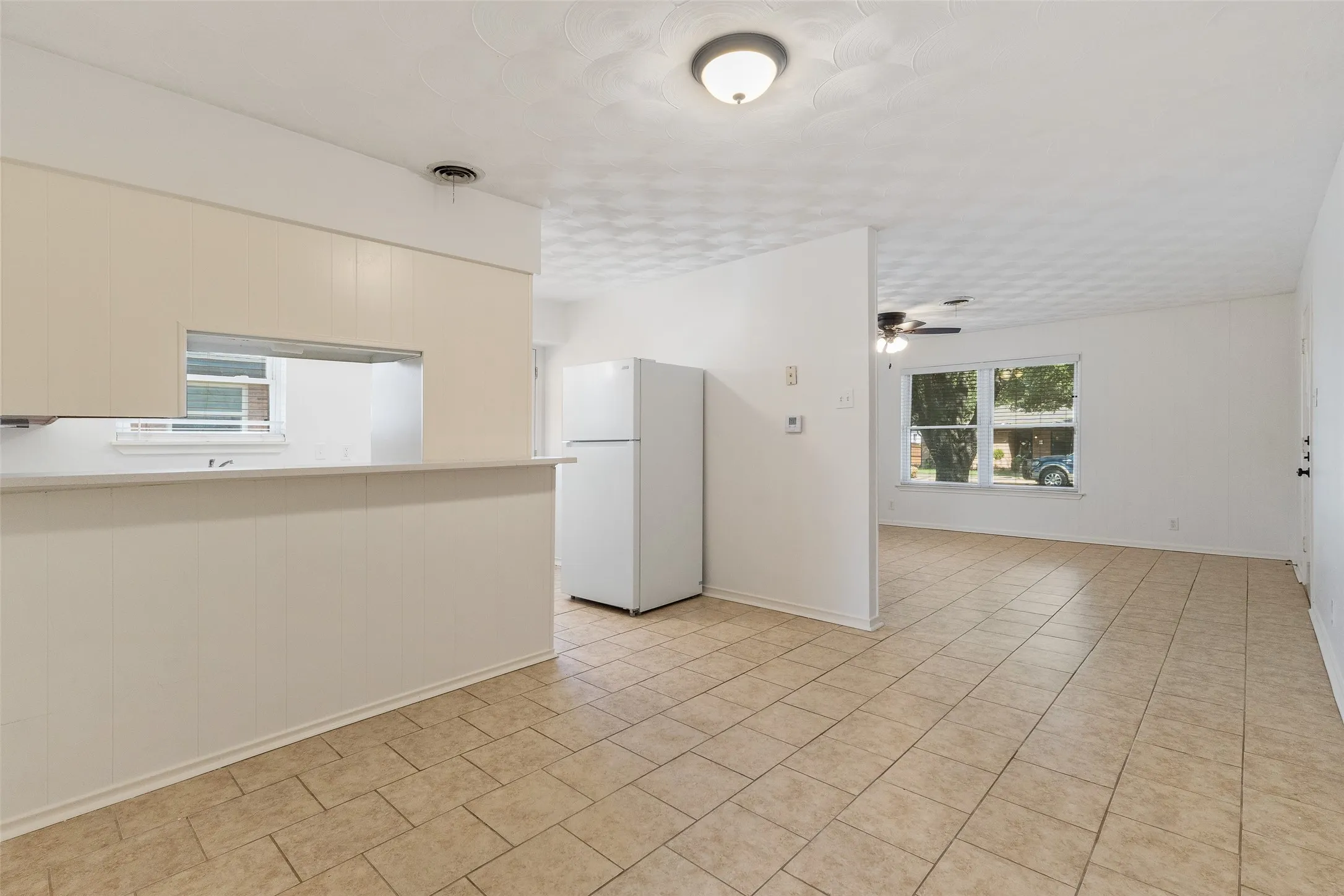 Kitchen area featuring a pass-through opening to the common space, neutral tile flooring, light-toned cabinetry, and a ceiling-mounted light fixture