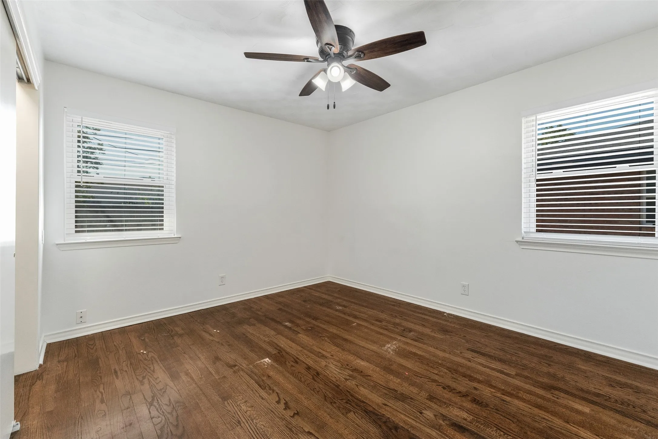 Wood-finish flooring, dual windows with horizontal blinds, and a central ceiling fan with integrated lighting