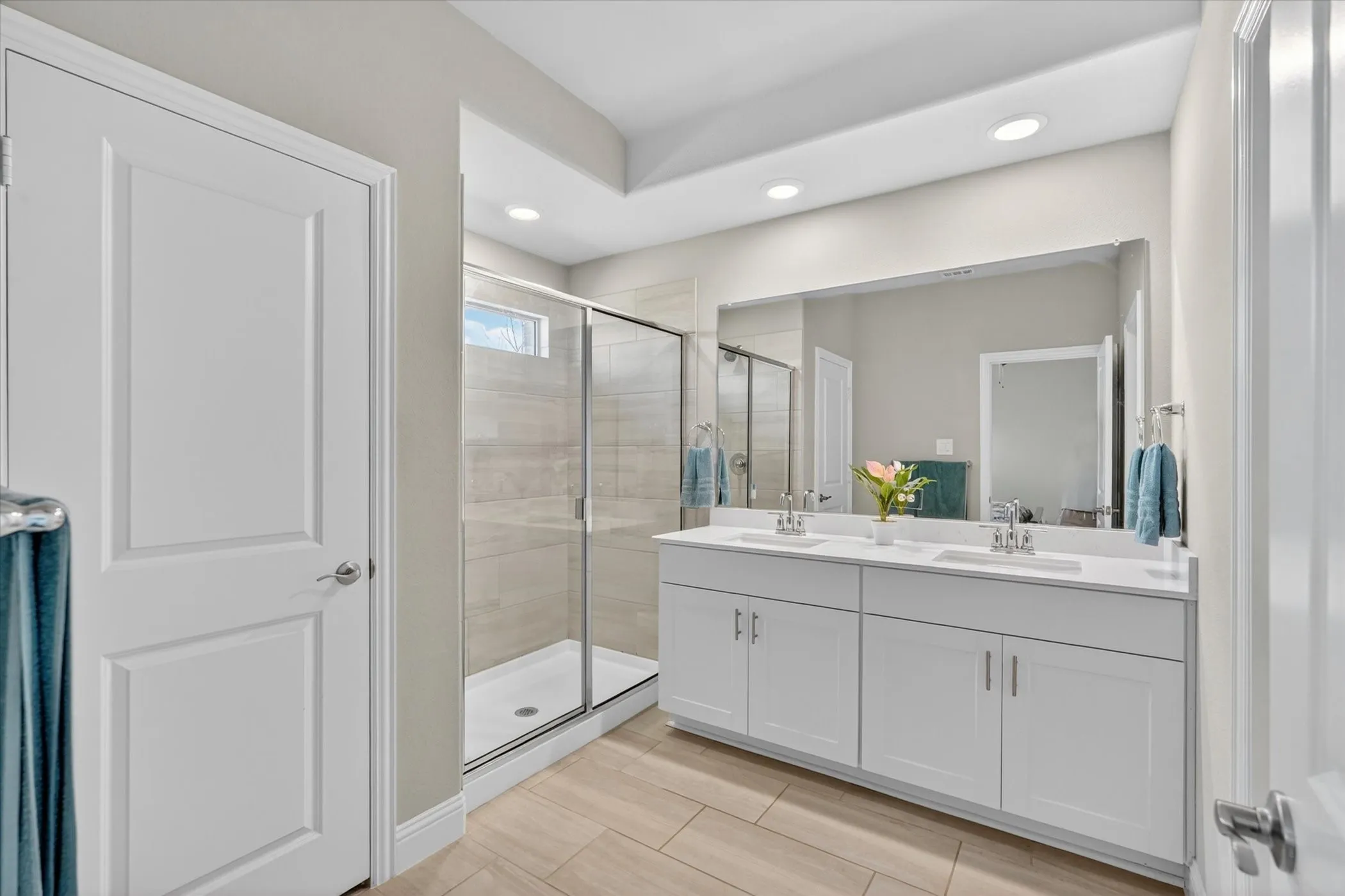 Bathroom featuring a dual vanity with white cabinetry and integrated sinks, a large frameless mirror, and a glass-enclosed shower with tile surround