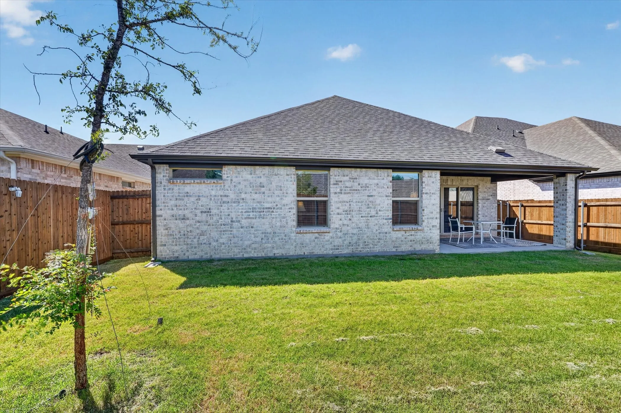 Expansive green lawn with a covered patio, featuring a brick exterior with multiple windows, a dark shingle roof, and wood fencing