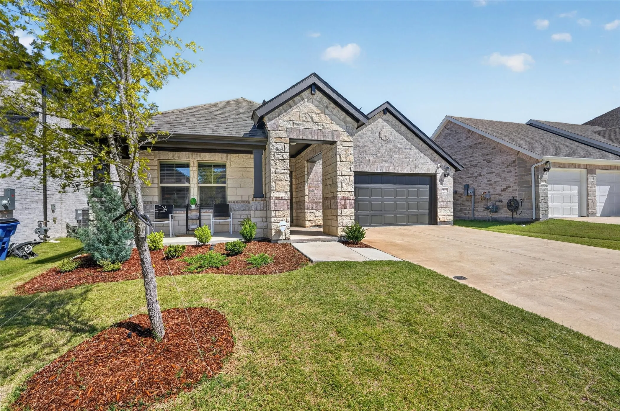 Single-story residence featuring a stone and brick exterior, an attached garage with a dark panel door, a covered porch, a concrete driveway, and landscaped front yard with mulch beds
