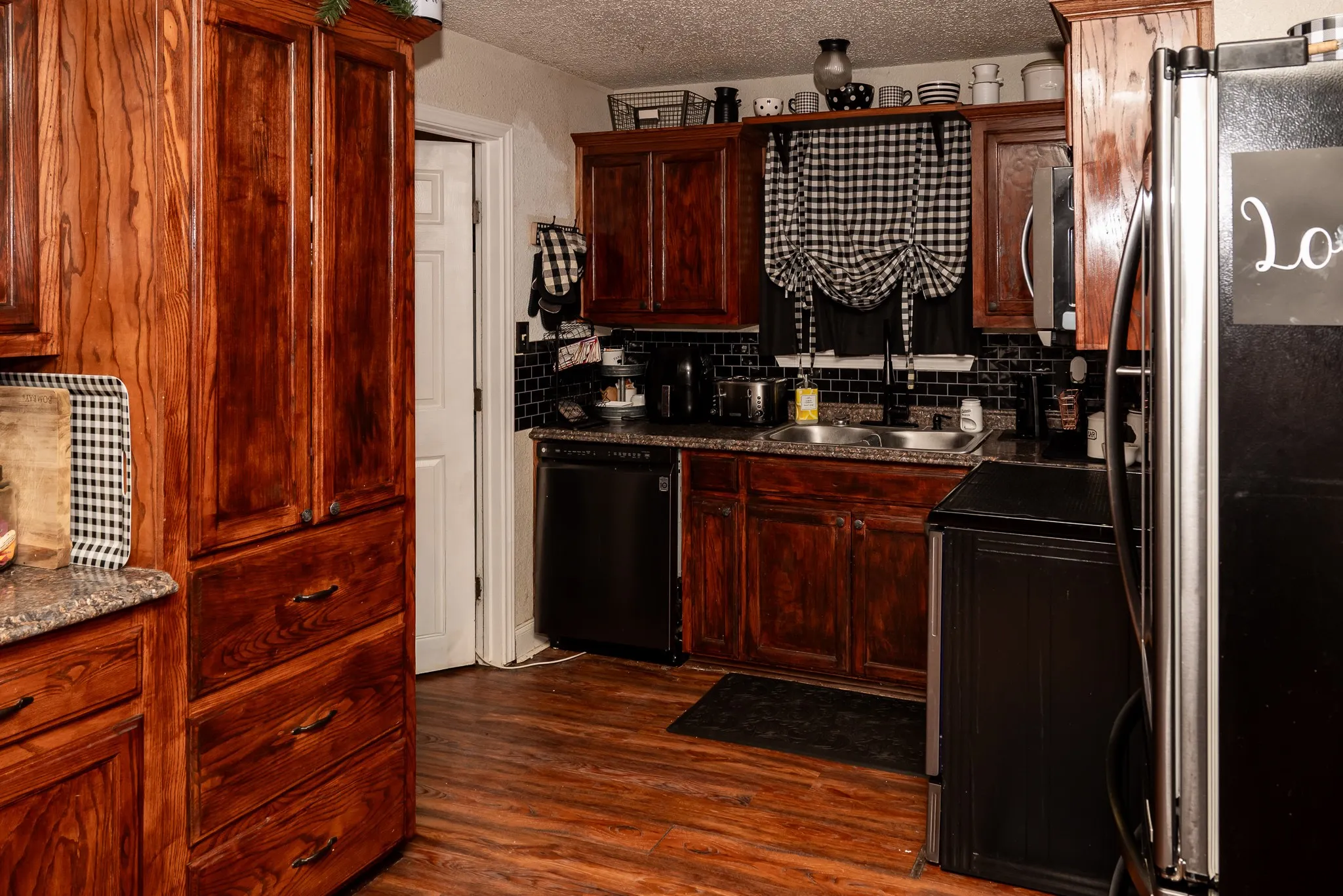 Kitchen featuring rich-toned wood cabinetry, black subway tile backsplash, stone-finish countertops, stainless steel sink with a high-arch faucet, and wood-finish flooring