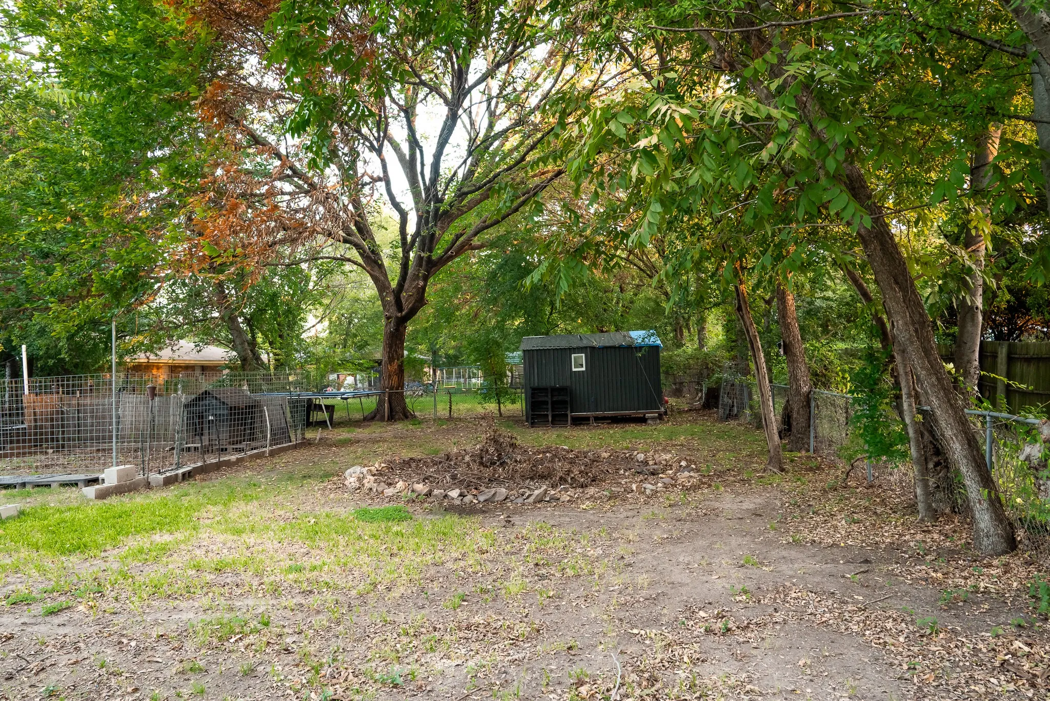 Expansive backyard featuring mature trees, a metal storage shed, a stone-bordered fire pit area, and perimeter chain-link fencing