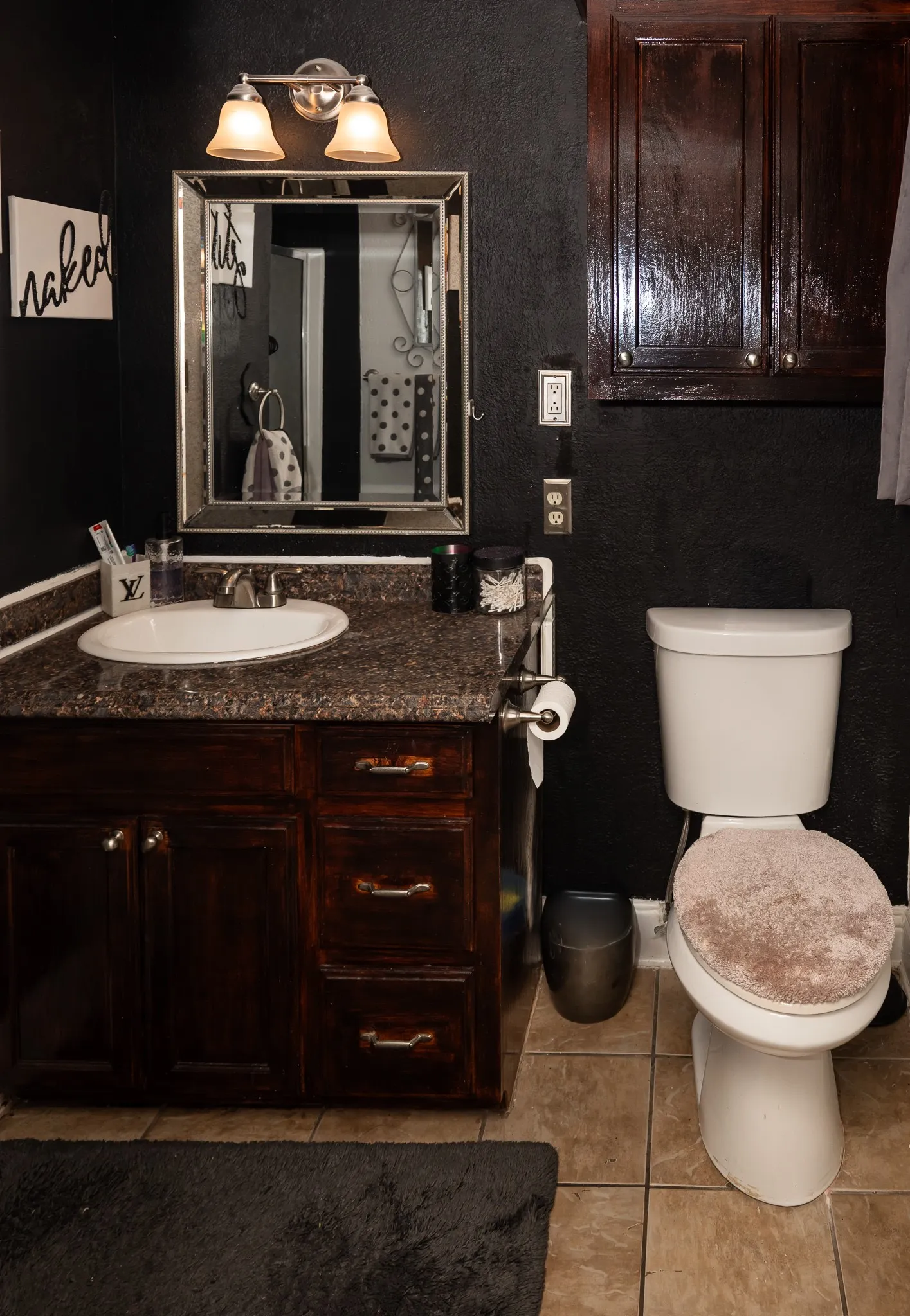 Bathroom vanity featuring stained wood cabinetry, stone-finish countertop with an integrated basin, wall-mounted dual-light fixture, framed mirror, and upper wall-mounted storage cabinet