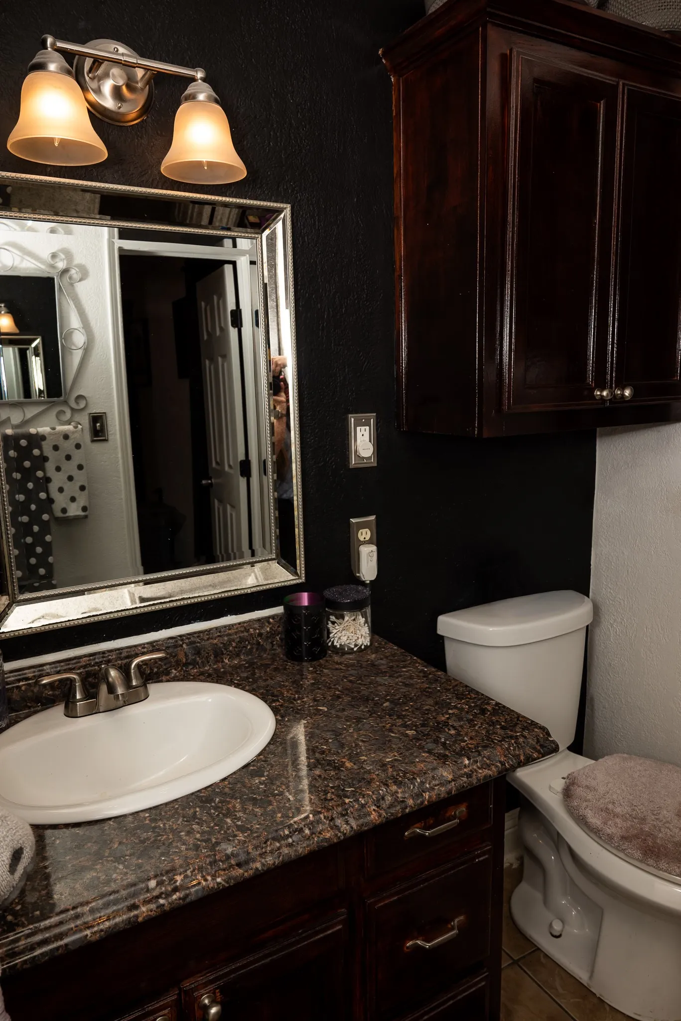 Dark-stained cabinetry, stone-surface countertop with an integrated basin, framed wall mirror, dual-shade vanity lighting, and built-in upper storage cabinet