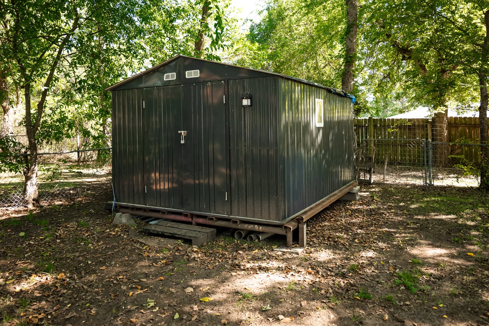 Detached storage structure featuring charcoal-colored corrugated metal siding, double swing doors, wall-mounted exterior lighting, and a raised metal frame foundation