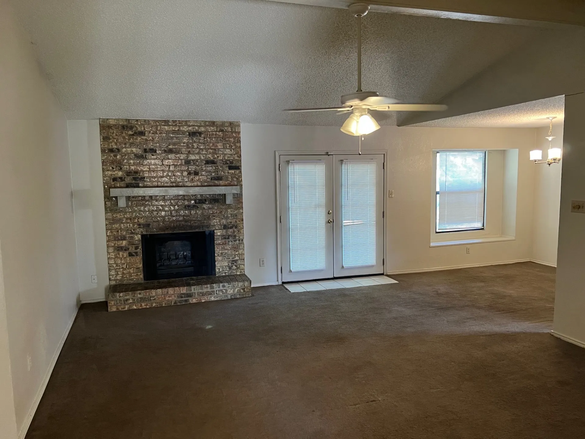 Vaulted ceiling, brick fireplace with hearth, ceiling fan with integrated lighting, glass-paneled double doors, and a window with a deep sill