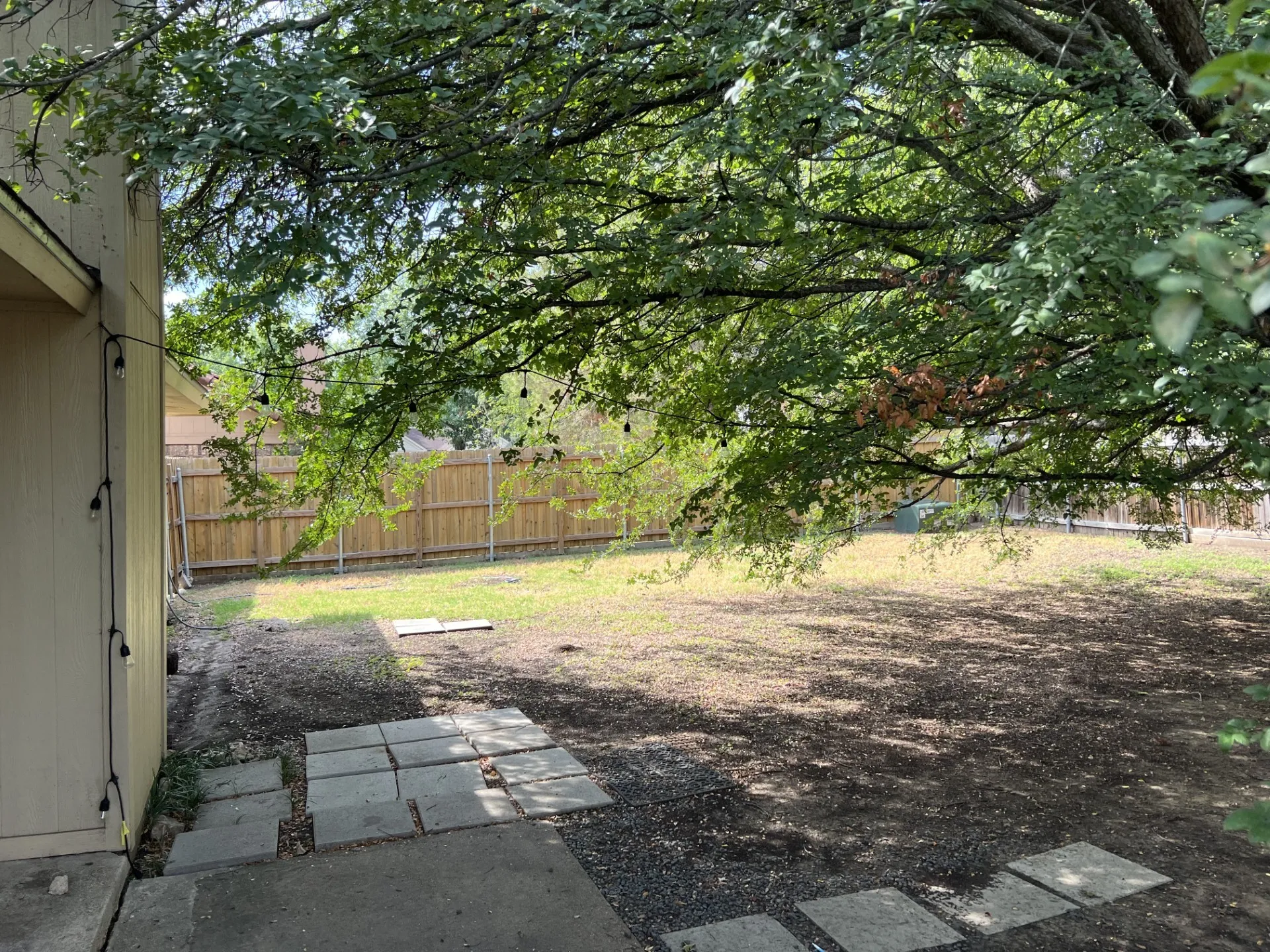 Expansive backyard featuring a natural ground cover, mature tree, concrete stepping stones, and a wooden fence
