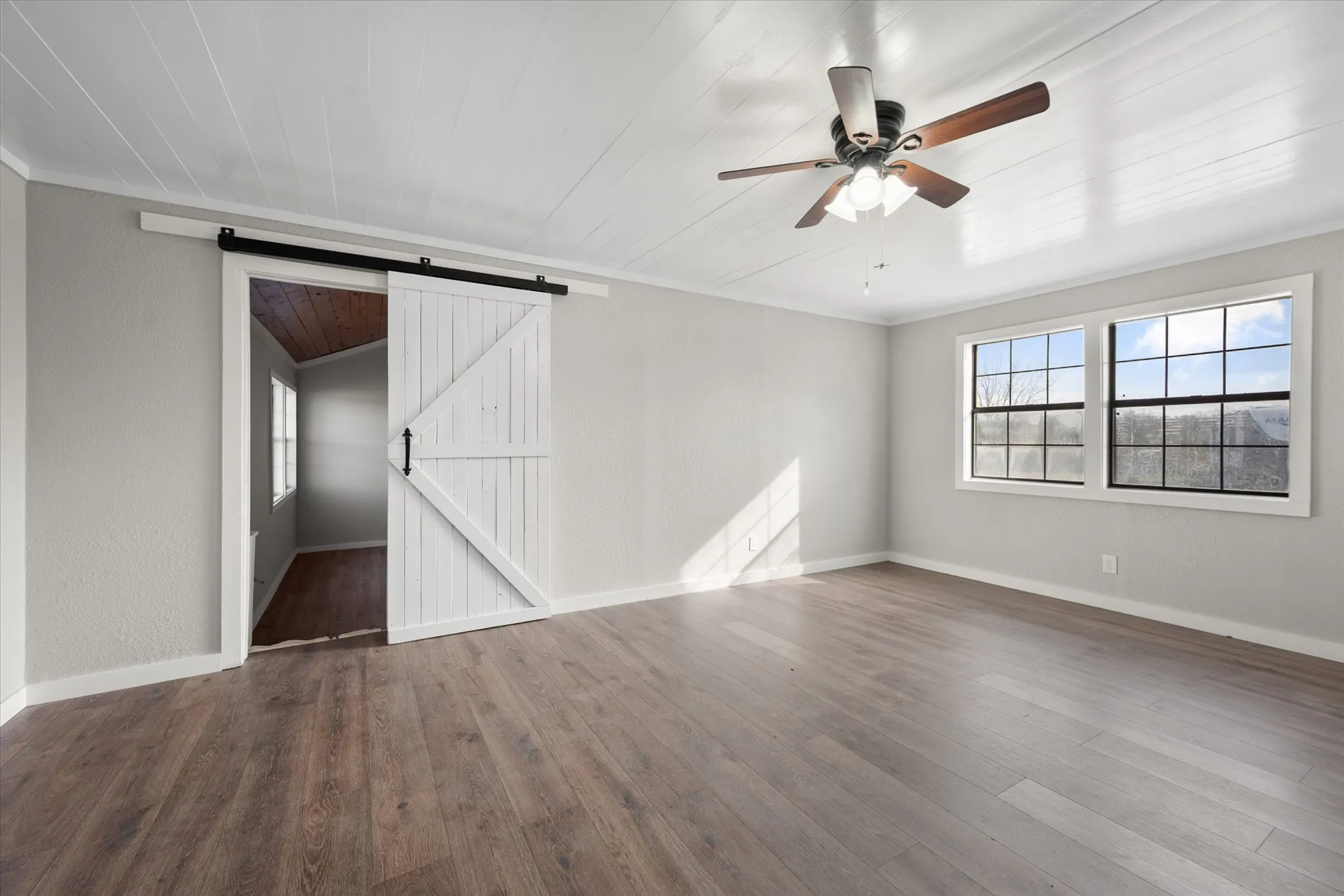 Wood-finish flooring, white shiplap ceiling, ceiling fan with light fixture, dual pane windows, and a white barn door