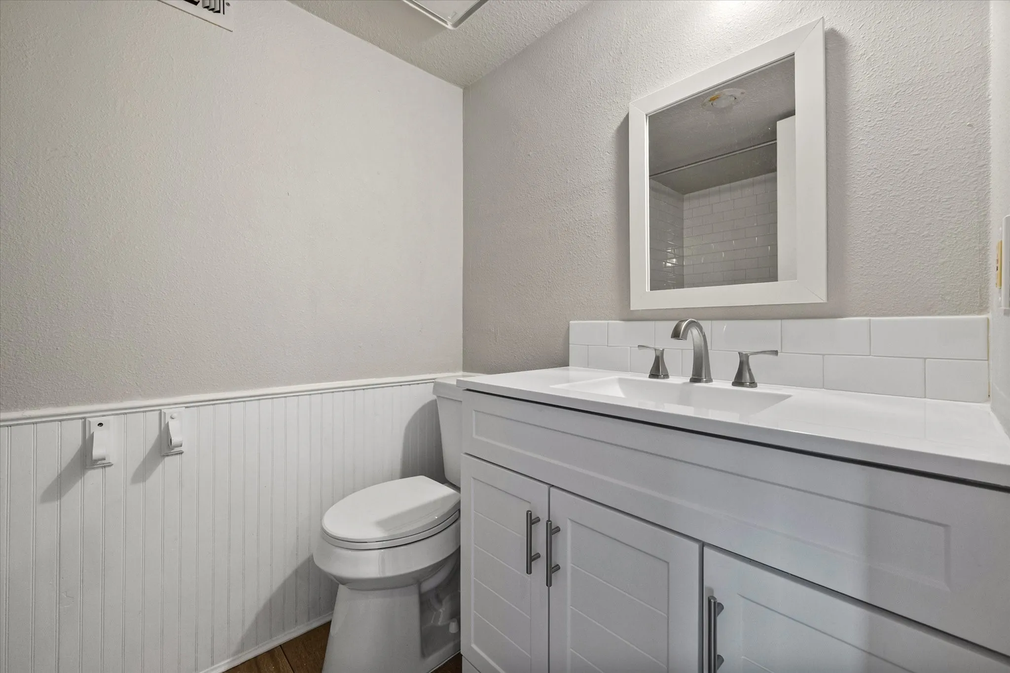 White vanity with integrated sink, brushed nickel faucet, white subway tile backsplash, white framed mirror, and wood-finish flooring