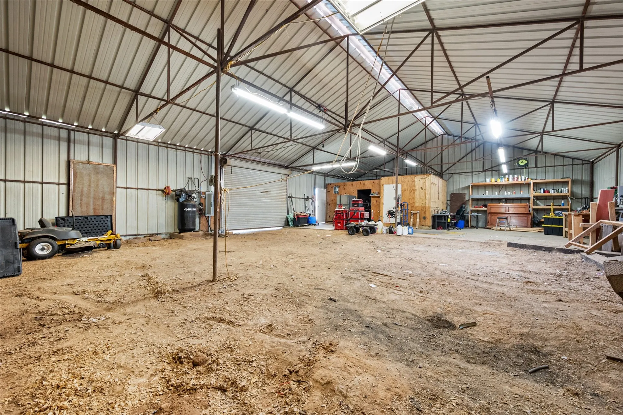 Large open warehouse space with corrugated metal walls and ceiling, exposed metal truss system, multiple skylights, and fluorescent strip lighting