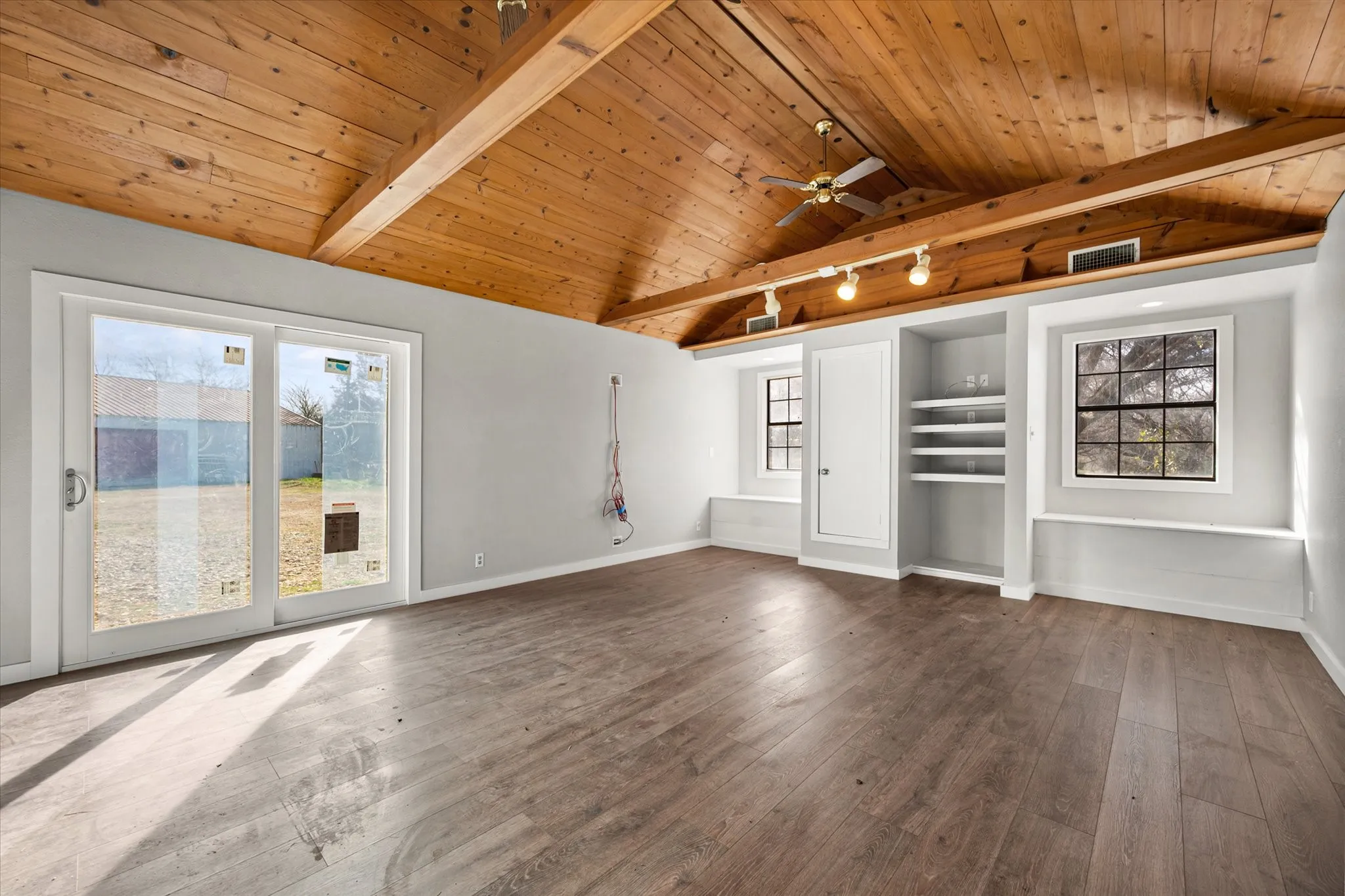 Vaulted wood-paneled ceiling with exposed beams, wood-finish flooring, built-in shelving, track lighting, and sliding glass doors