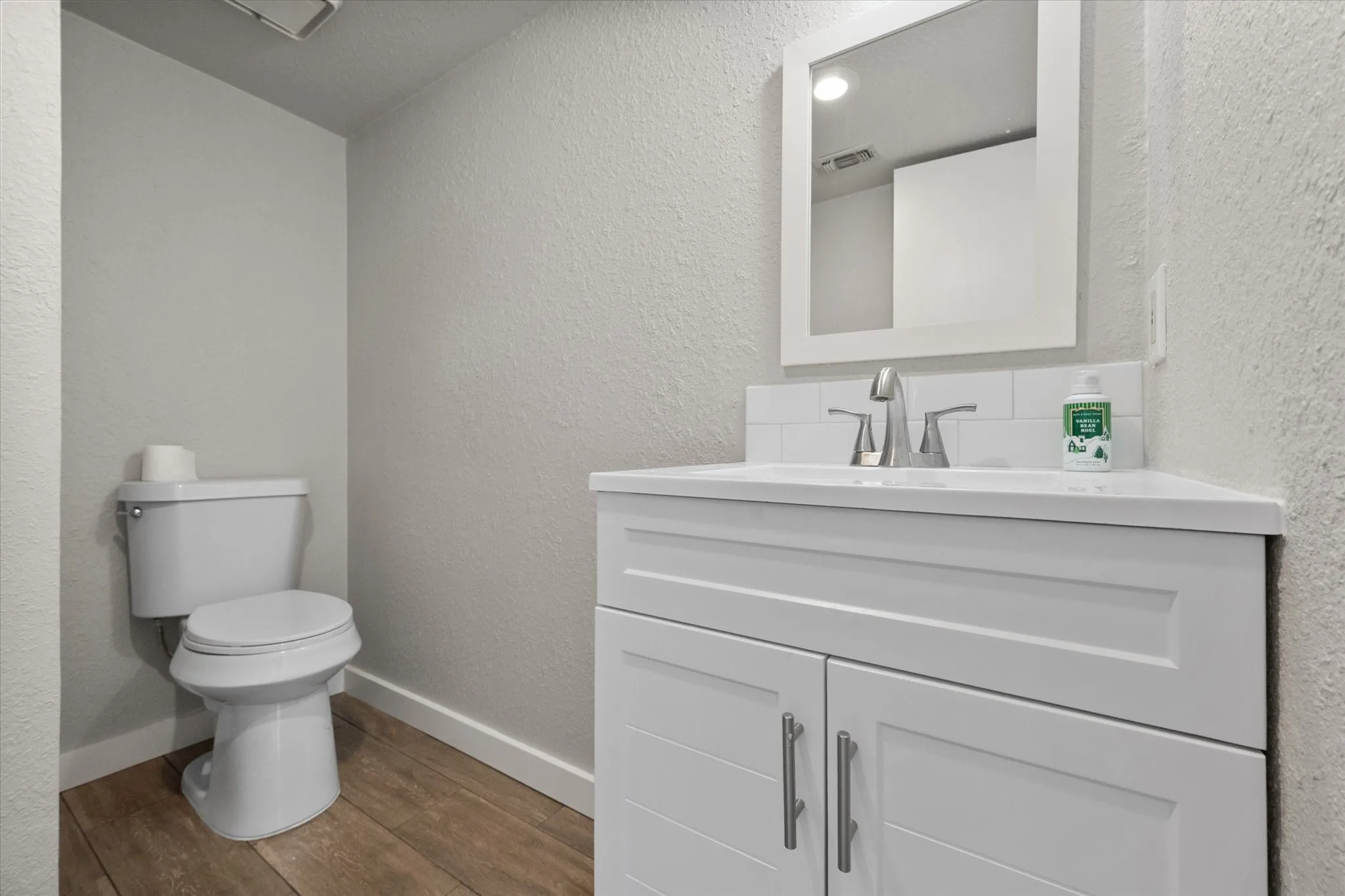 White vanity with brushed nickel hardware, white rectangular sink, white subway tile backsplash, and wood-finish flooring