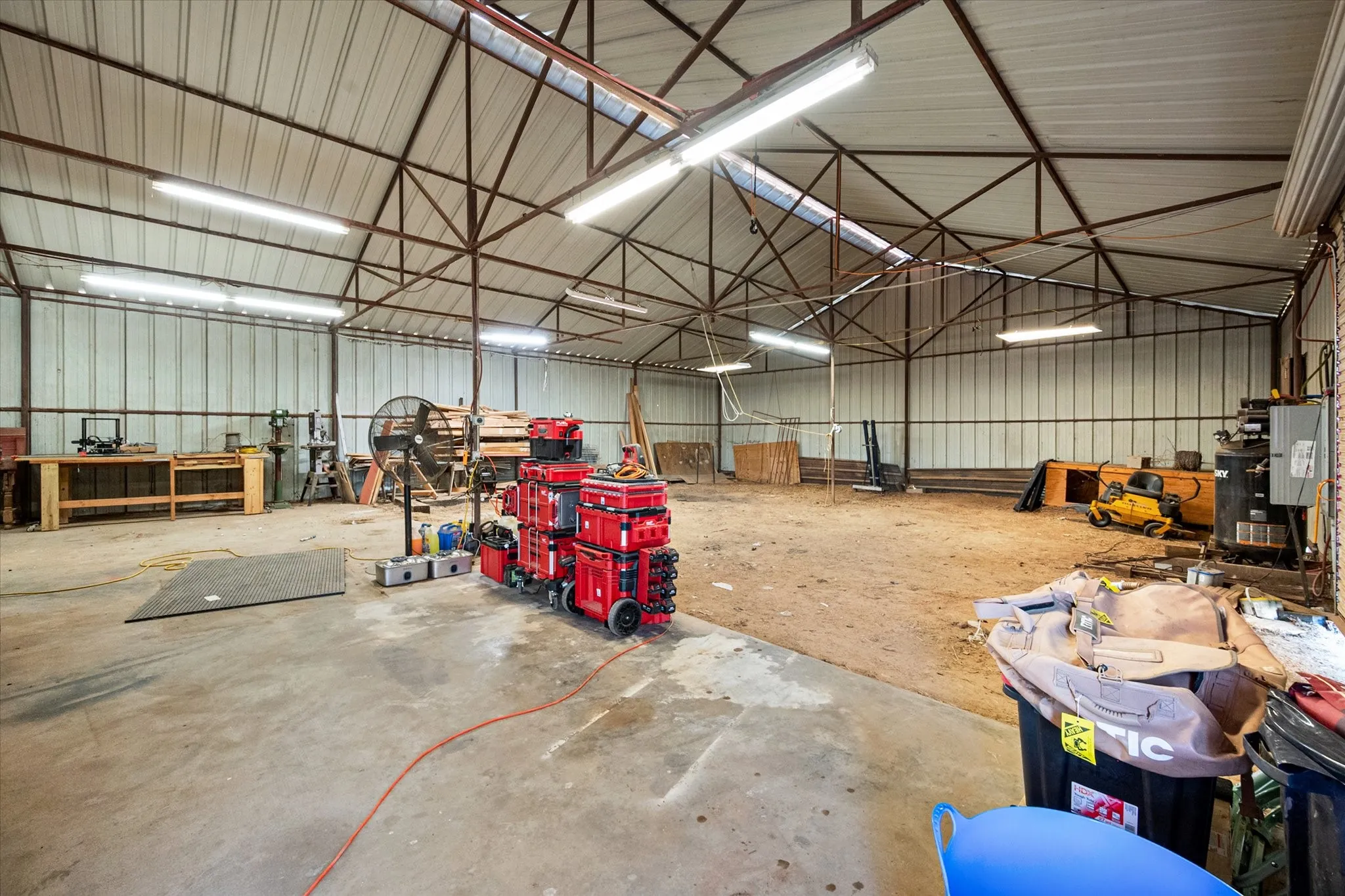 Warehouse structure featuring a metal roof, exposed truss system, concrete flooring, corrugated metal walls, and overhead fluorescent lighting