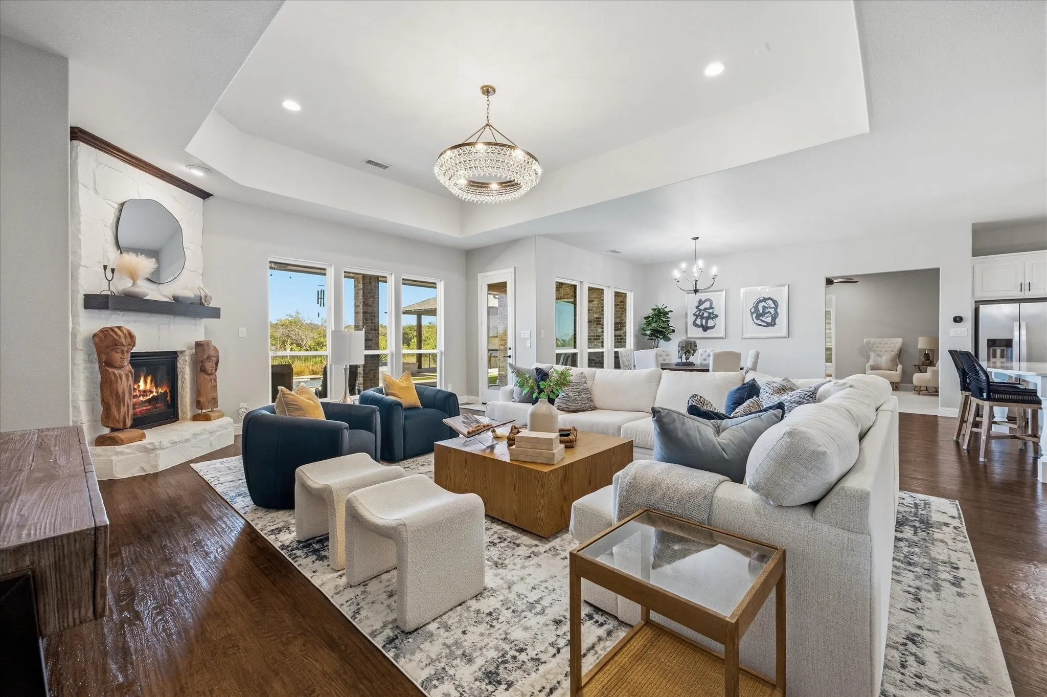 Expansive living area featuring wood-finish flooring, a white brick fireplace with a dark mantel, a tray ceiling, and large windows