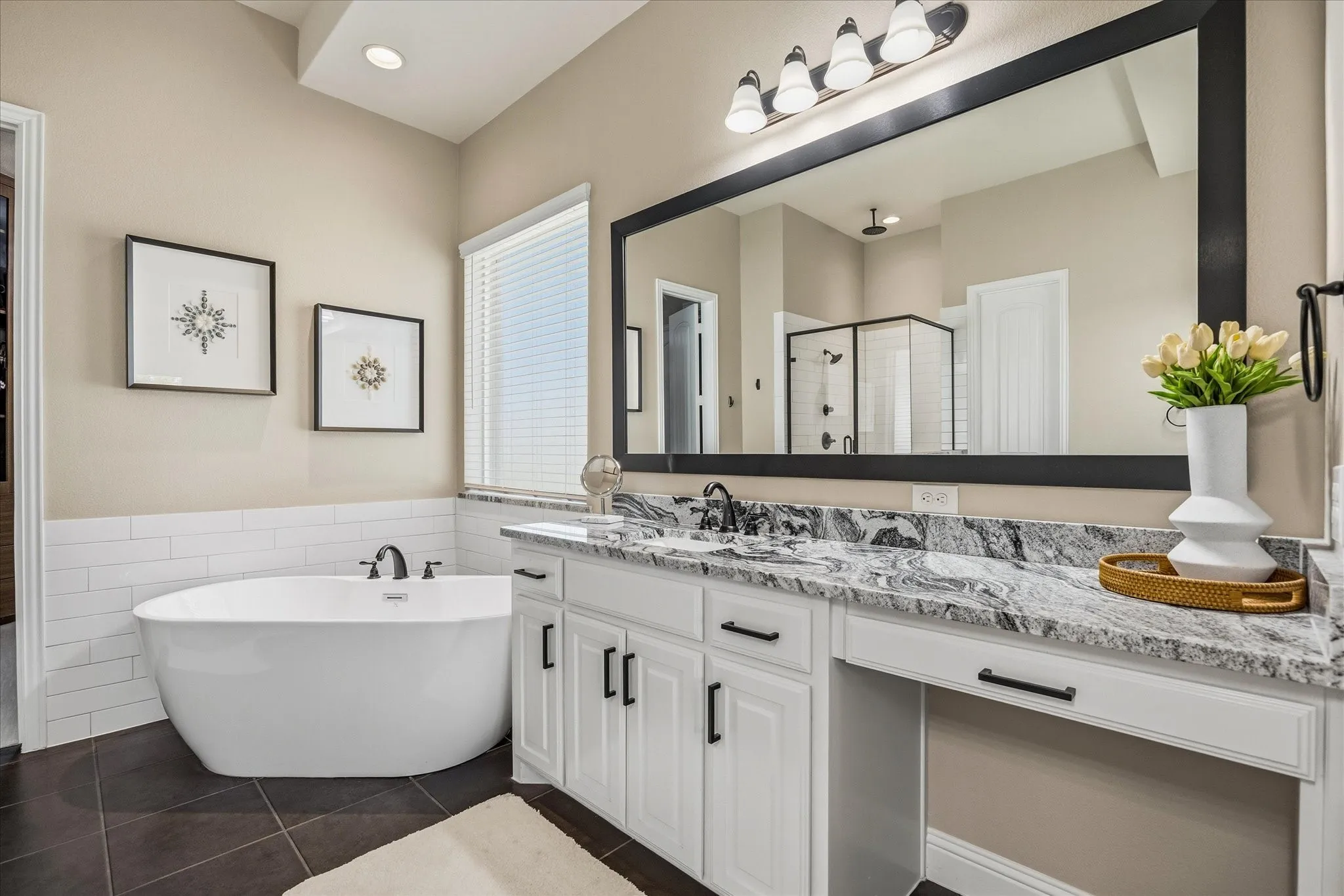 Freestanding soaking tub with dark bronze fixtures, white subway tile surround, double vanity with granite countertop, dark bronze hardware, and a large framed mirror