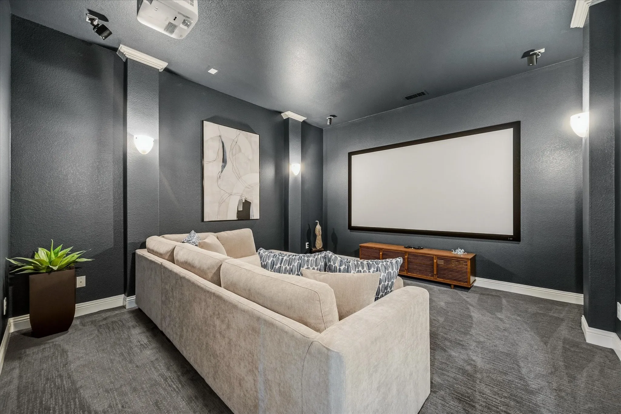Dedicated media room featuring a large projection screen, mounted projector, recessed lighting, wall sconces, and a dark carpeted floor