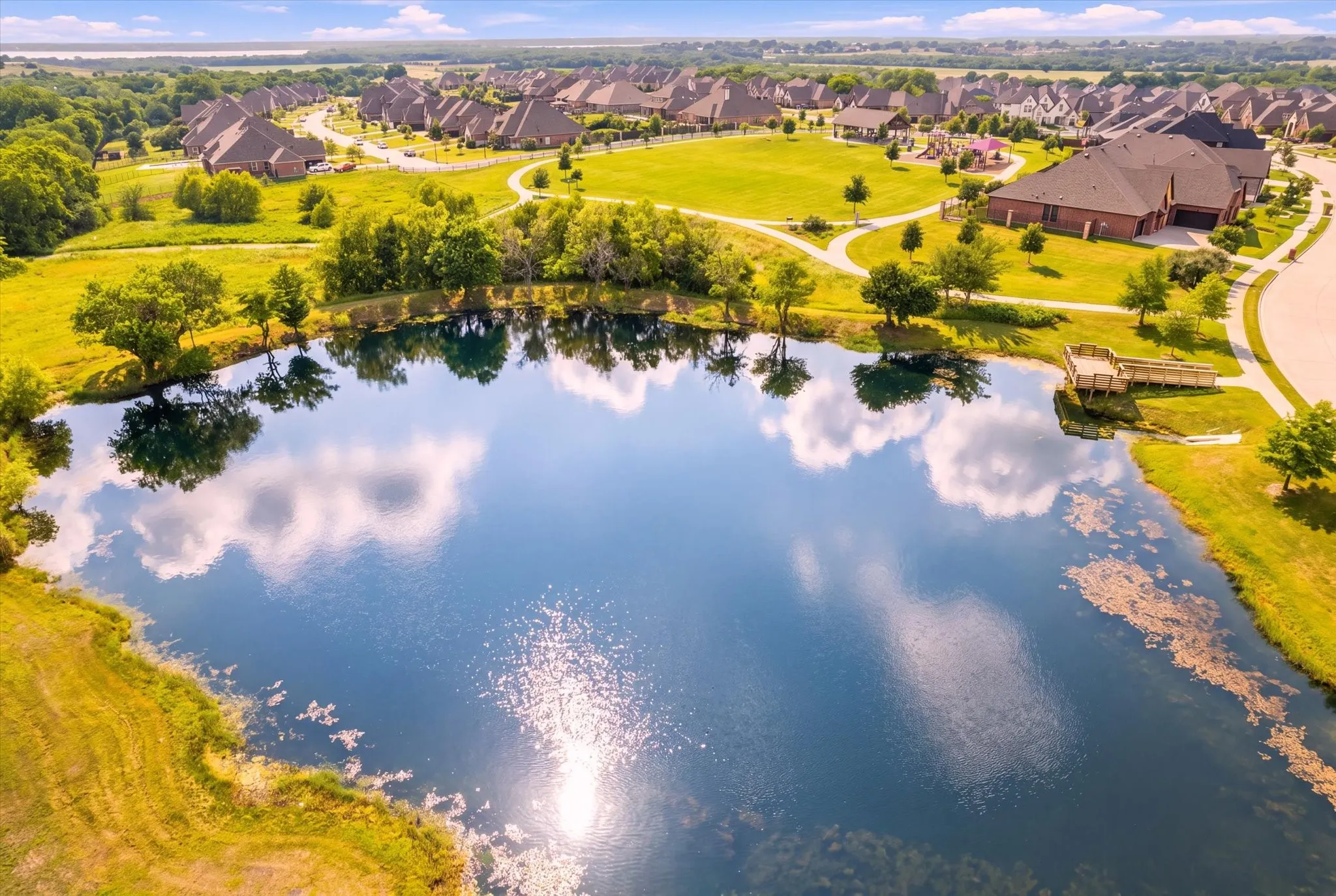 Residential community featuring a prominent pond with a wooden dock, surrounded by green spaces and paved pathways