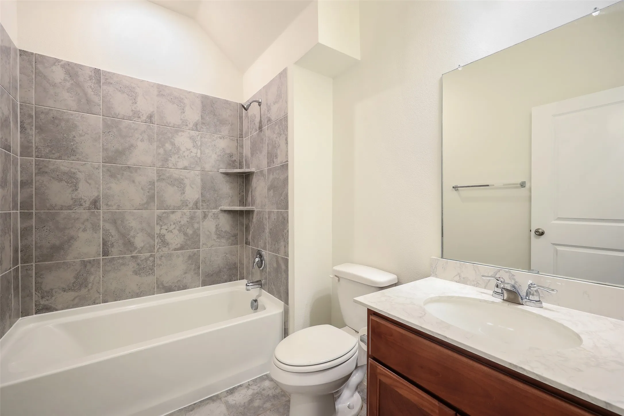 Full bathroom featuring a tub-shower combination with floor-to-ceiling tile surround, built-in shelving, wood-finish vanity with a stone-top integrated sink, and an expansive wall-mounted mirror