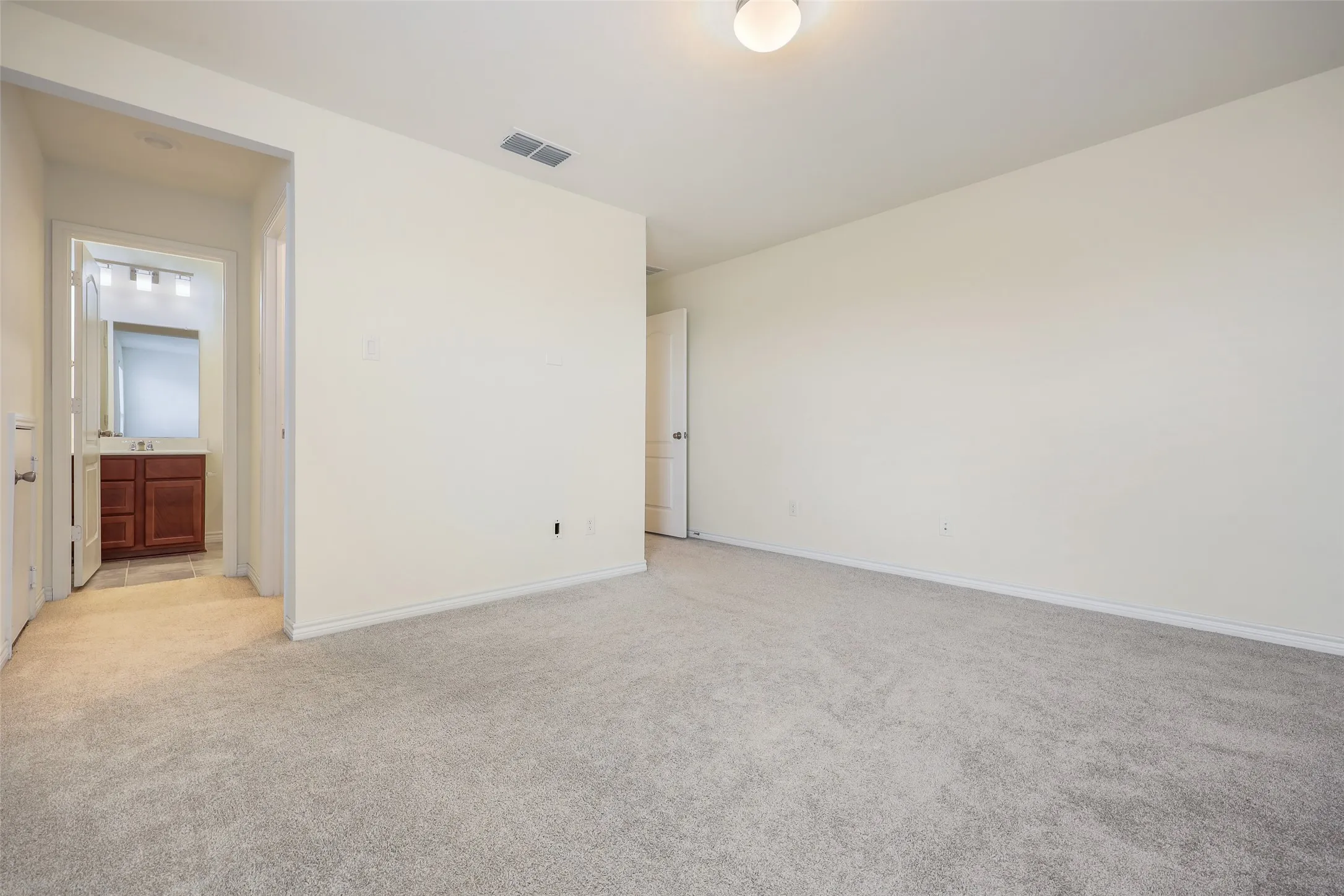 Neutral-toned interior space featuring wall-to-wall carpeting, flush-mount ceiling lighting, and a doorway revealing a bathroom with wood-finish cabinetry and a vanity mirror