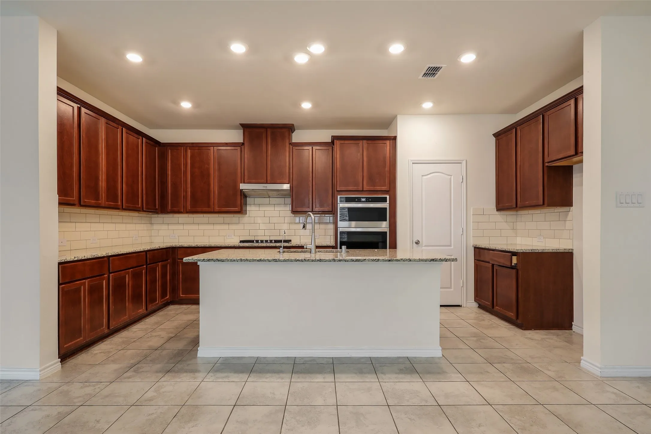 Kitchen featuring stained wood cabinetry, stone-surface countertops, subway tile backsplash, center island with sink, and stainless steel double wall ovens