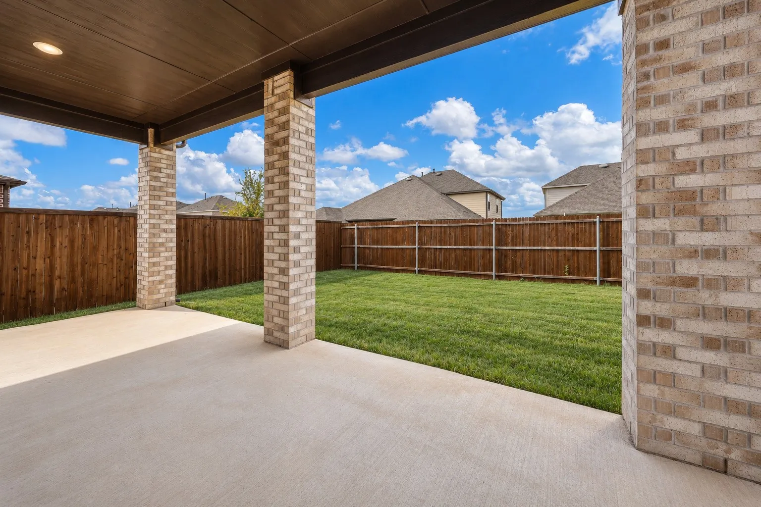 Virtually improved image. Covered patio featuring a concrete floor, recessed ceiling lighting, and brick support columns, opening to a fenced backyard with a lawn