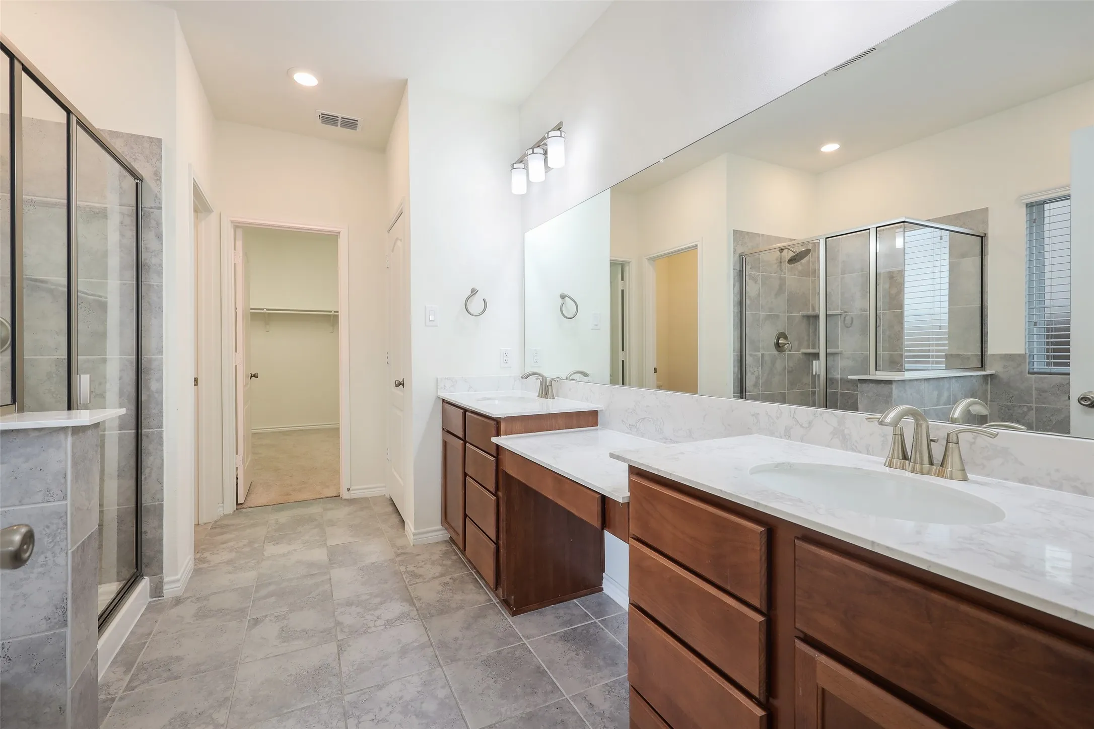 Dual-sink vanity with stone-finish countertops, wood cabinetry, glass-enclosed shower with tile surround, and full-width vanity mirror