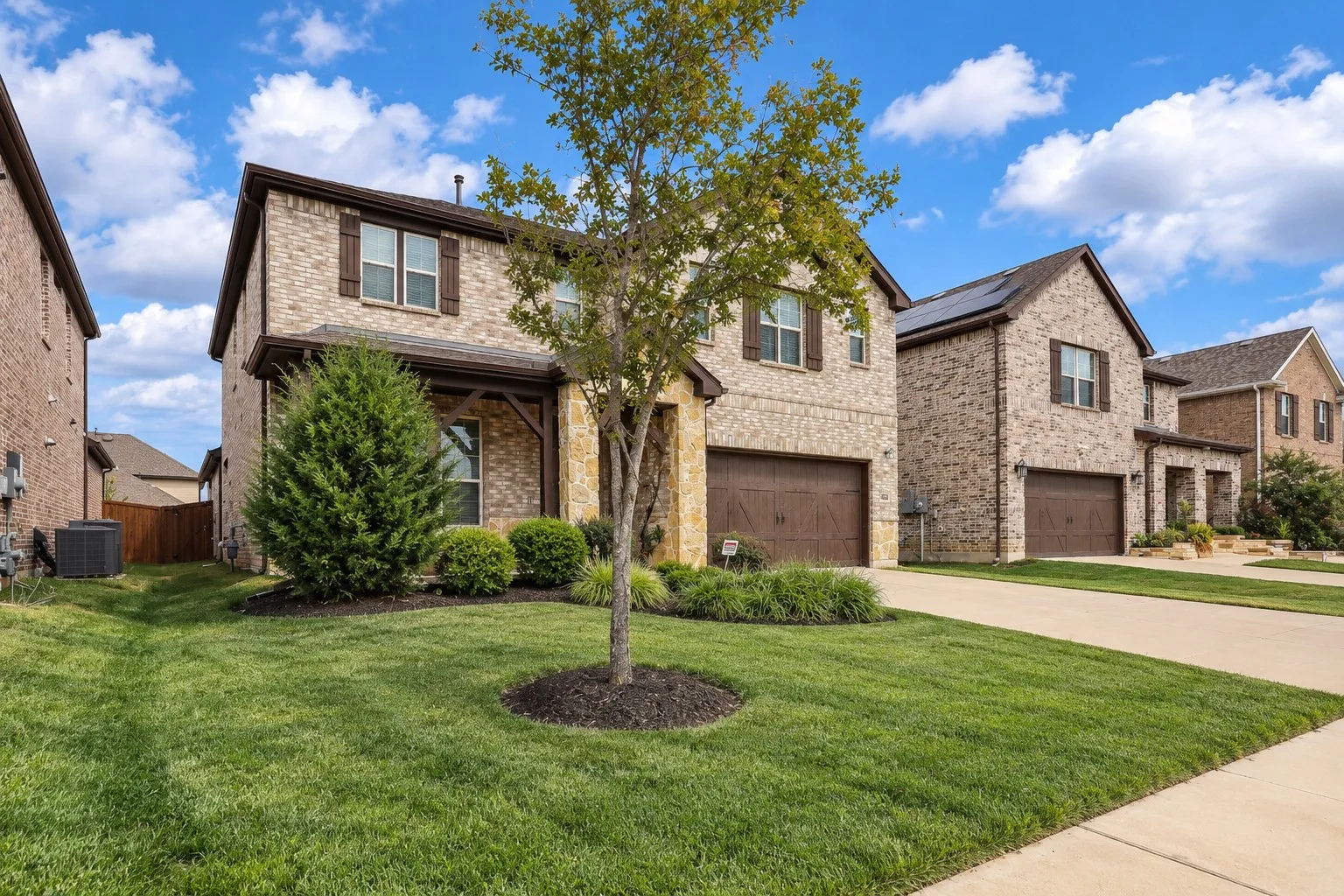 Virtually improved image. Two-story residence featuring a mixed brick and stone facade, covered front entryway with timber-style bracing, attached two-car garage, and manicured front lawn with mature landscaping