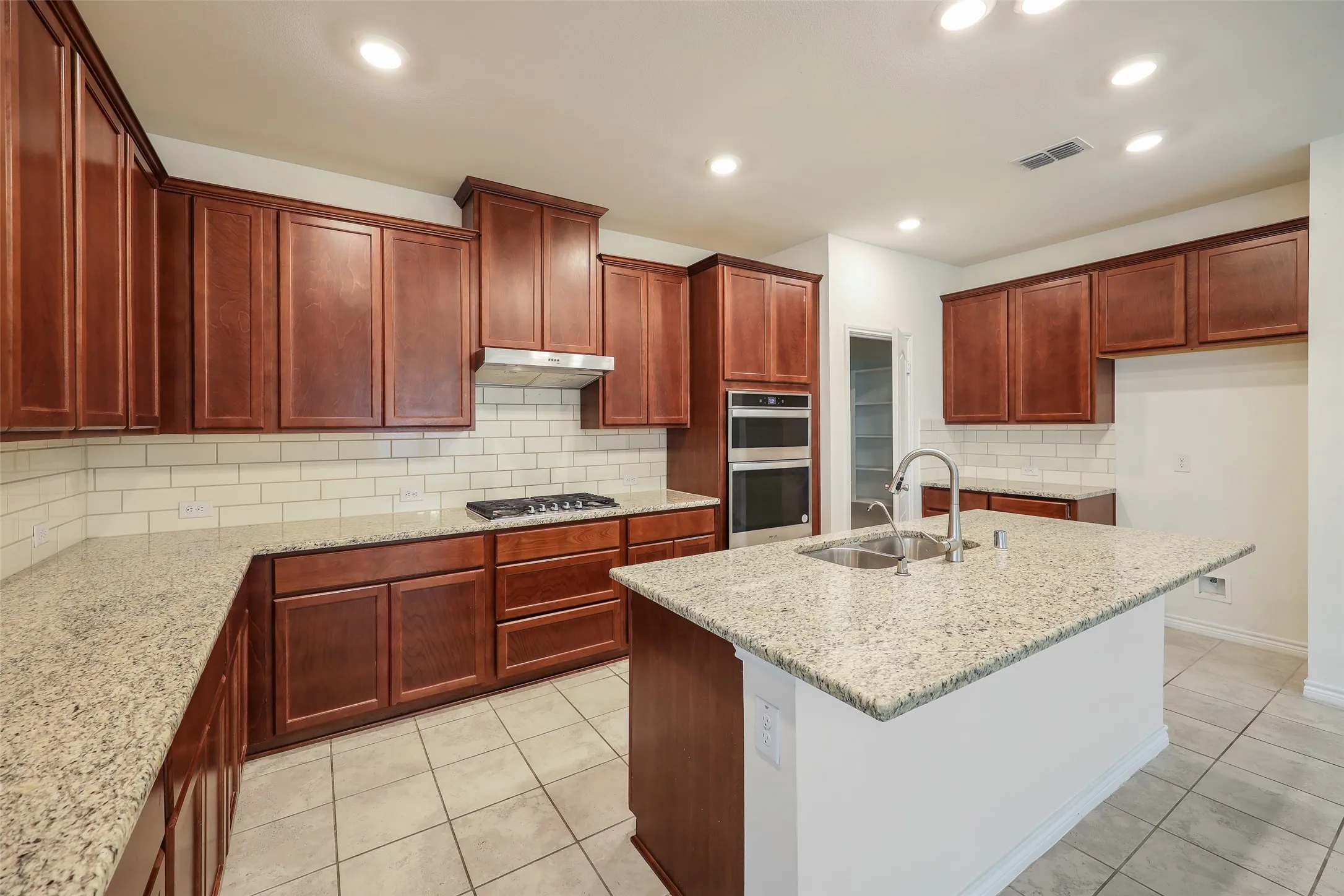 Kitchen featuring rich wood cabinetry, stone-surface countertops, subway tile backsplash, stainless steel double-wall ovens, and a central island with an integrated sink