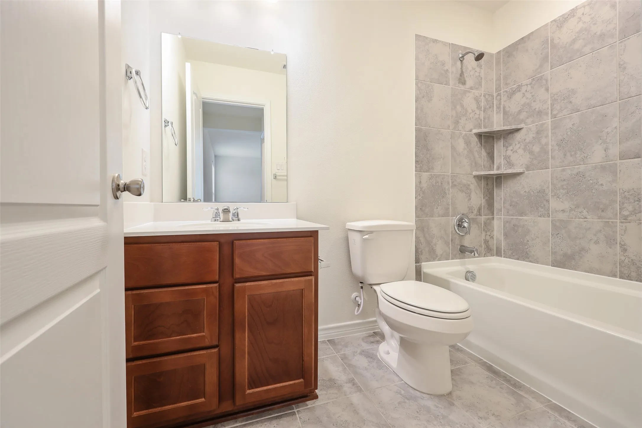 Bathroom featuring a wood-finish vanity with a white countertop, large wall-mounted mirror, tub-shower combination with gray tile surround, and built-in shelving