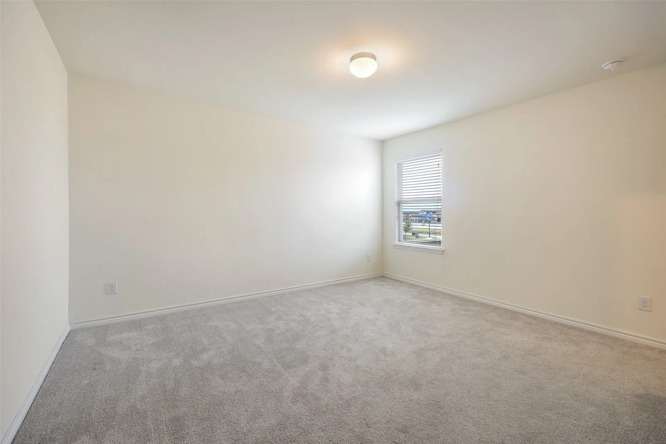 Neutral-toned interior room featuring wall-to-wall carpeting, a single-pane window with integrated blinds, and a flush-mount ceiling light fixture