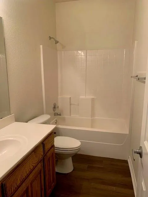 Bathroom featuring a wood-finish vanity with a white countertop, an integrated sink basin, a white tub-shower combination with surround, and wood-finish flooring