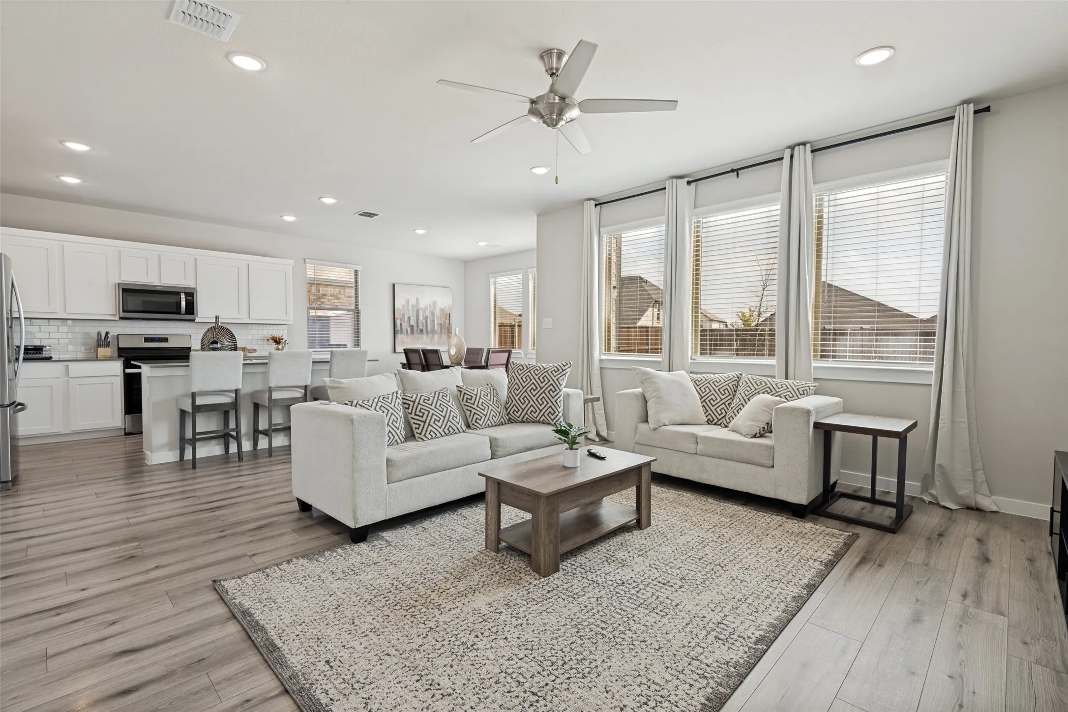 Open-concept main room featuring wood-finish flooring, recessed lighting, a brushed-nickel ceiling fan, and large windows with horizontal blinds