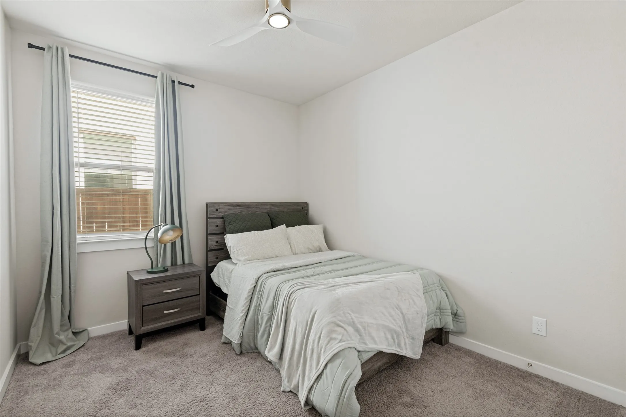 Neutral-toned interior featuring wall-to-wall carpeting, window with horizontal blinds, and a ceiling-mounted light fixture with fan blades