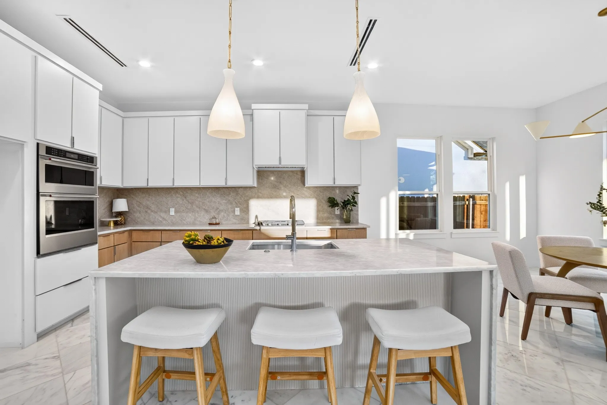 Kitchen featuring an oversized central island with stone-surface countertop, white cabinetry with wood-finish lower cupboards, stone-tiled backsplash, stainless steel double wall ovens, and pendant lighting