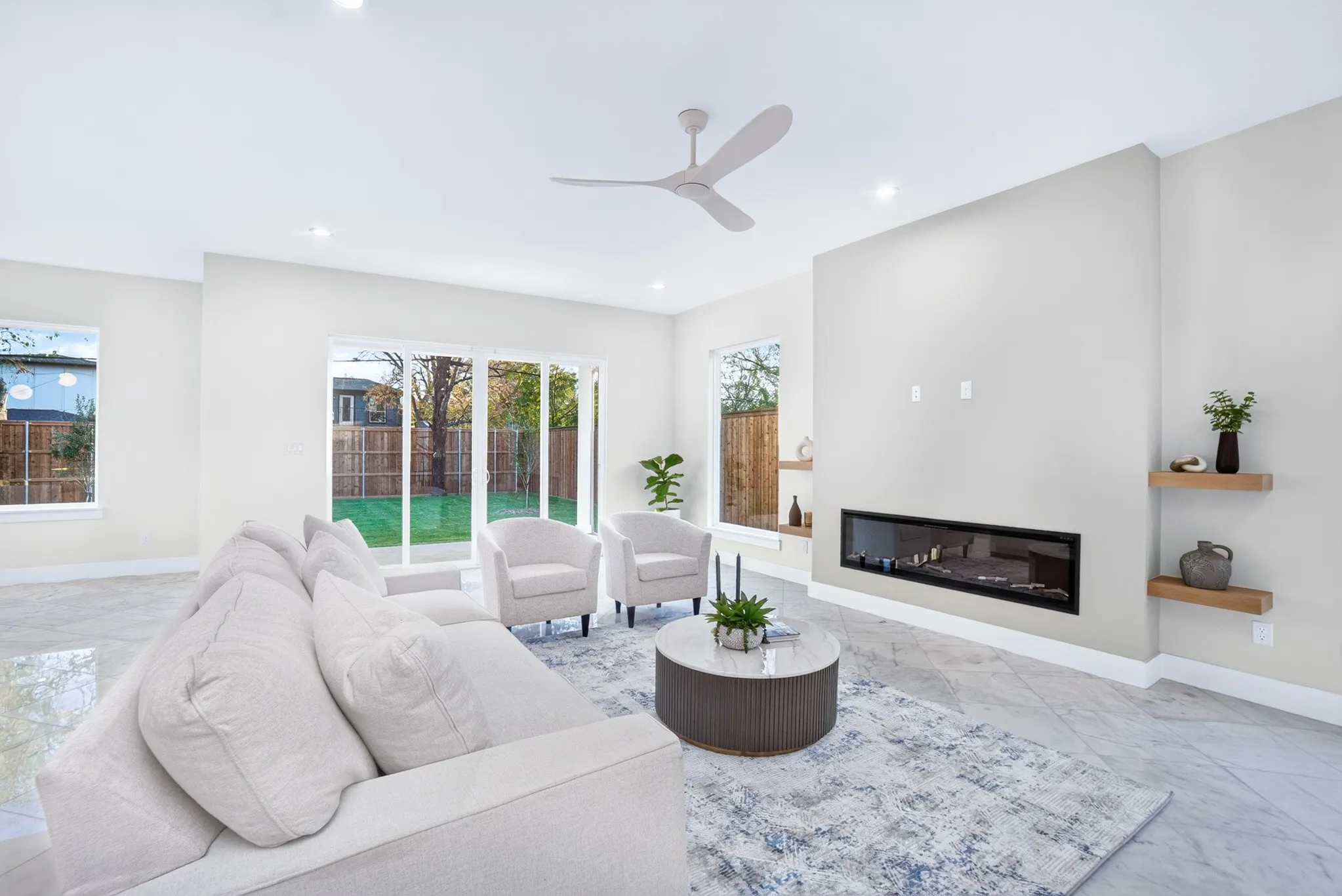 Living area featuring large-format tile flooring, a horizontal linear fireplace, wood-finish floating shelving, multi-panel sliding glass doors, and a modern ceiling fan
