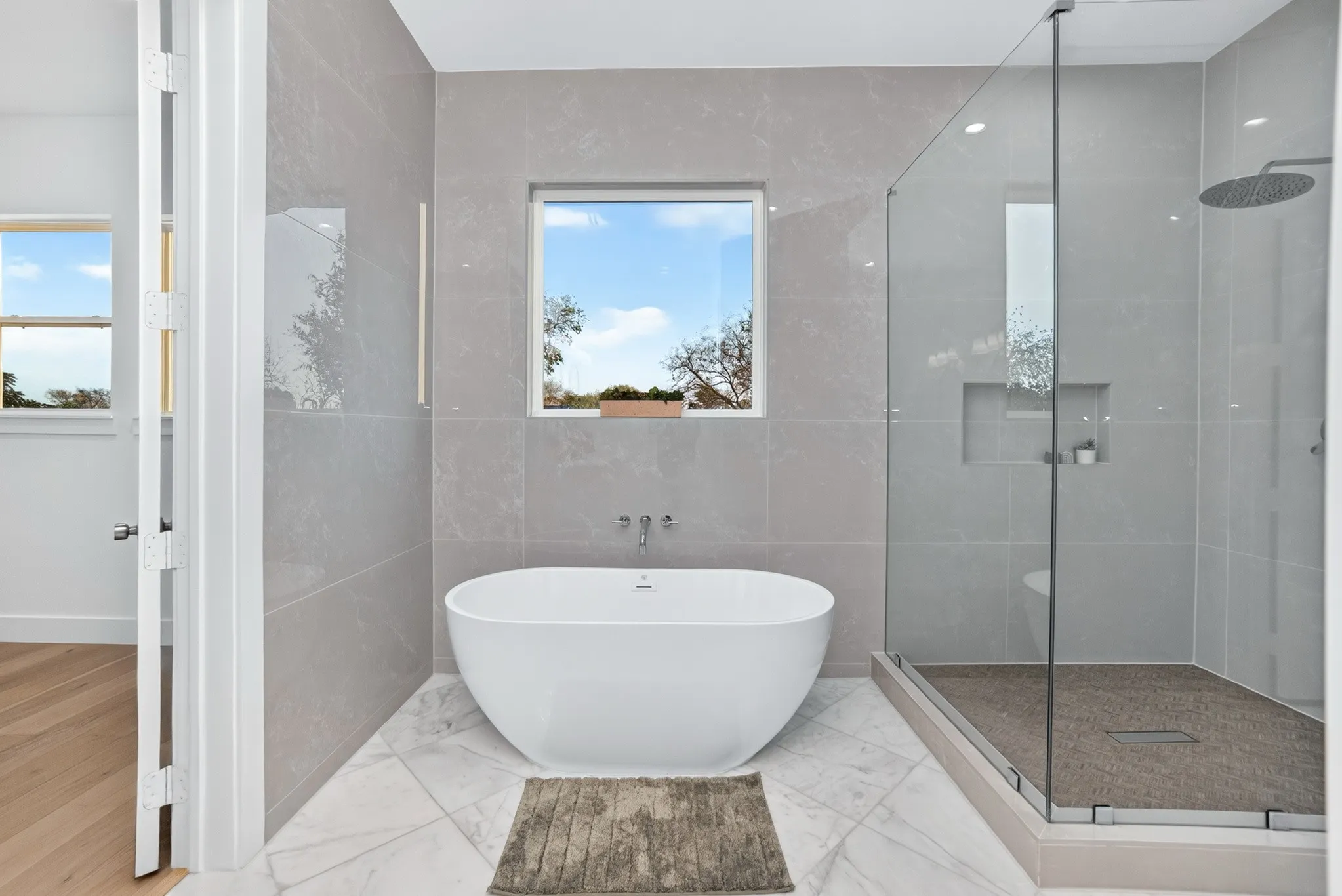 Freestanding soaking tub, walk-in glass-enclosed shower with rain-style showerhead, large-format wall tiling, recessed shower niche, and tile flooring