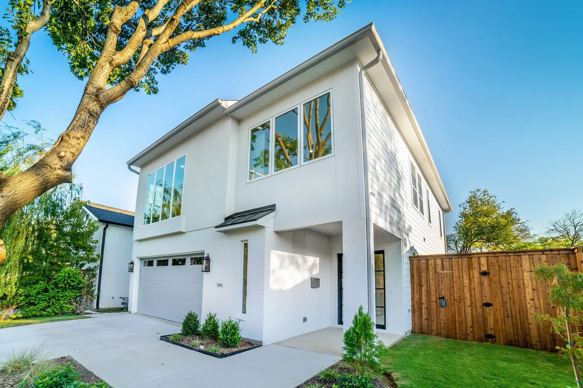 Two-story contemporary exterior featuring white masonry and horizontal siding, large-format windows, a concrete driveway, and a wooden privacy fence