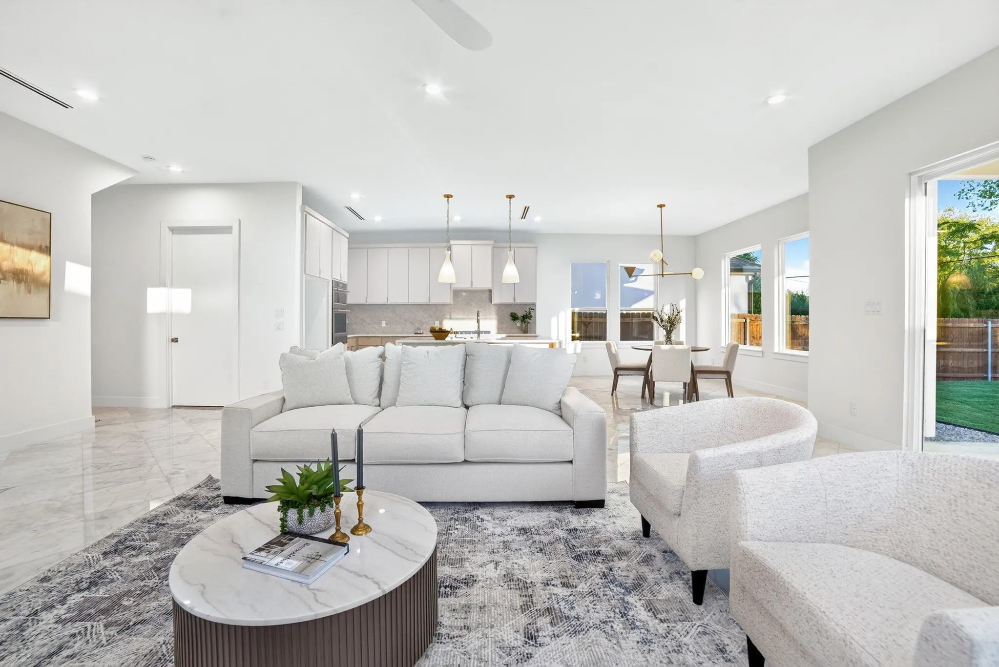 Open-concept interior featuring light-toned stone-finish tile flooring, recessed ceiling lighting, a contemporary kitchen with white cabinetry, and large windows providing natural light