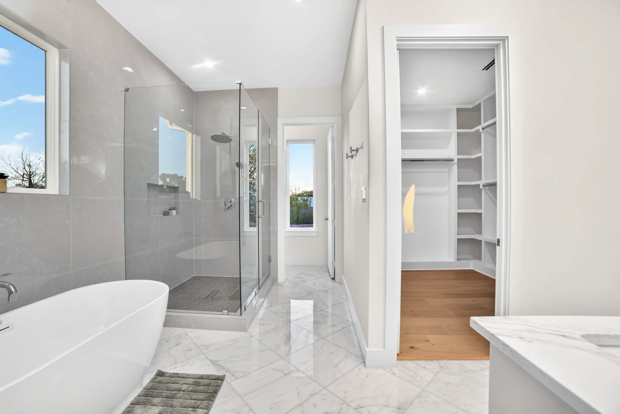 Freestanding soaking tub, glass-enclosed shower with tiled walls and recessed shelving, stone-finish tile flooring, and walk-in closet featuring built-in shelving and wood-finish flooring