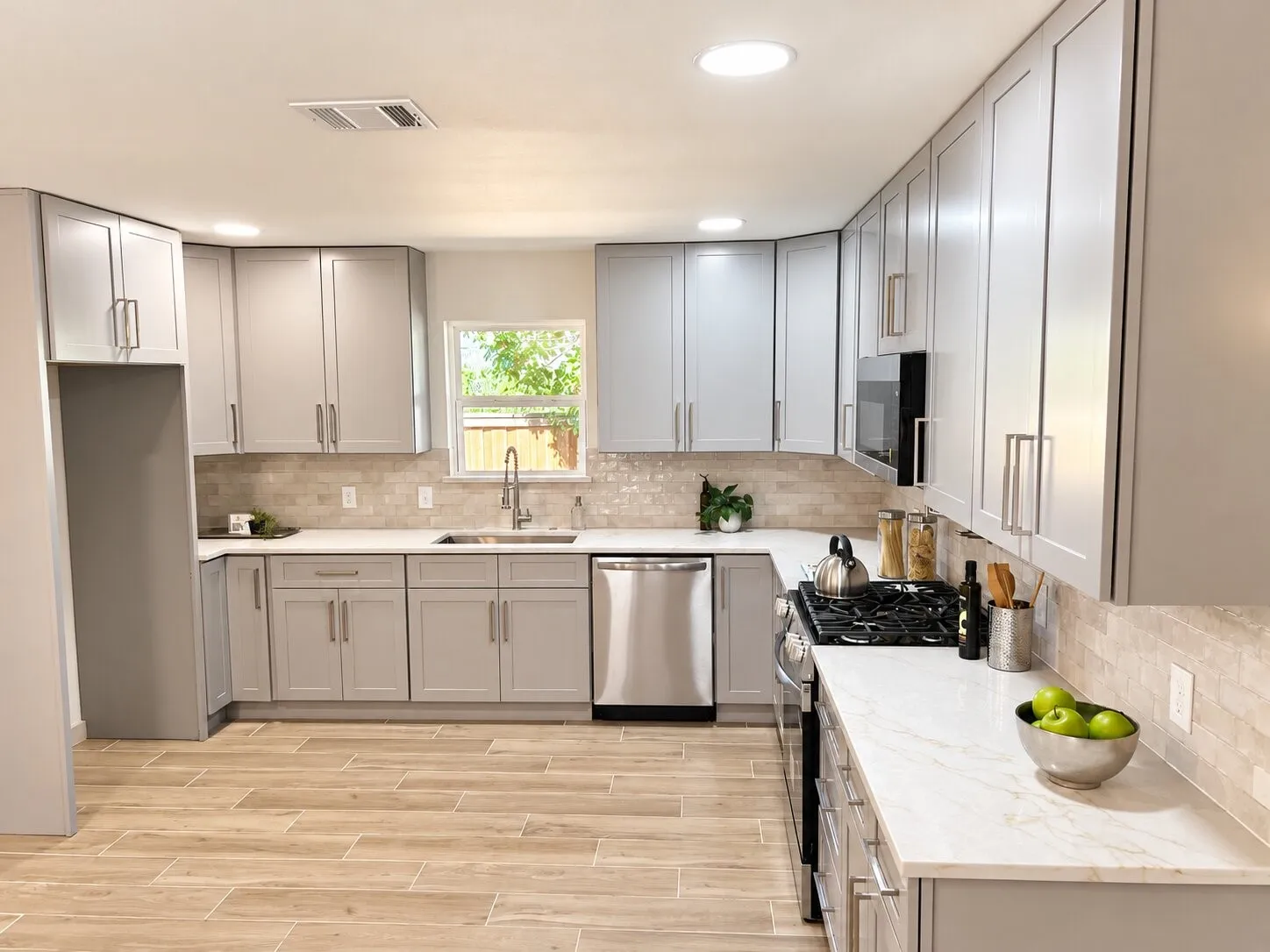 Kitchen featuring light grey cabinetry with brushed nickel hardware, white stone-finish countertops, subway-style tiled backsplash, stainless steel appliances, and wood-finish tile flooring