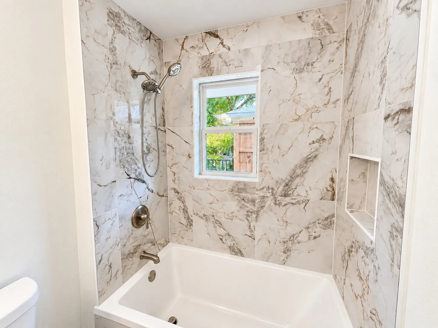 Bathtub surround featuring large-format stone-patterned tile, integrated recessed shelving, chrome handheld shower fixture, and a central window